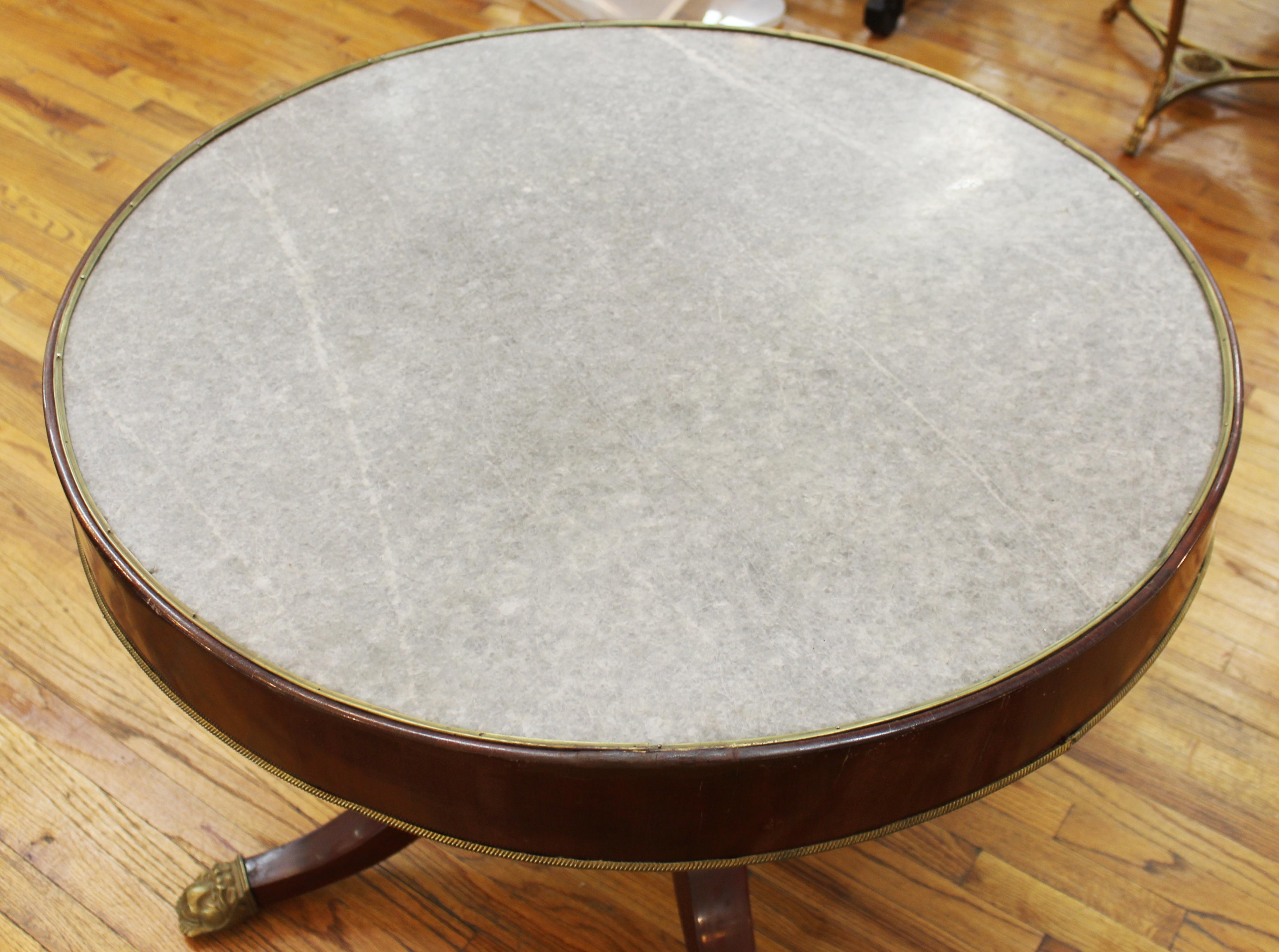 Regency style mahogany center table, the top with gilt metal banding and inset with grey marble risen above birdcage-form parcel ebonized columns supported by four curved legs terminating on brass lion-motif paw feet. Measures: 29” height x 37.75”