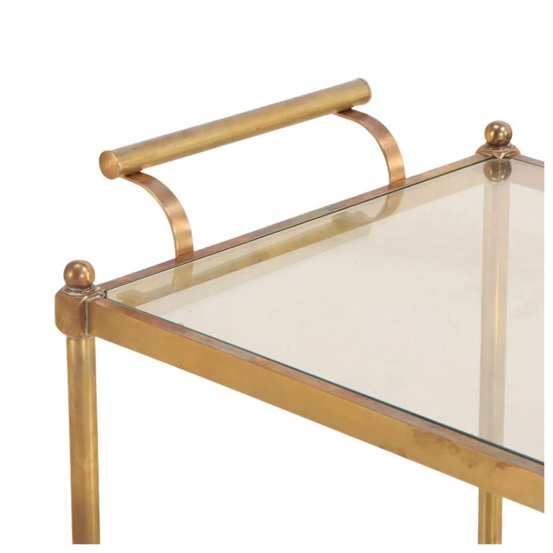 Questo sofisticato carrello da bar a due livelli in ottone e vetro, risalente al 1970 circa, incarna lo stile Regency con il suo design raffinato e la sua eleganza funzionale. La struttura in ottone lucido presenta graziose maniglie ricurve e