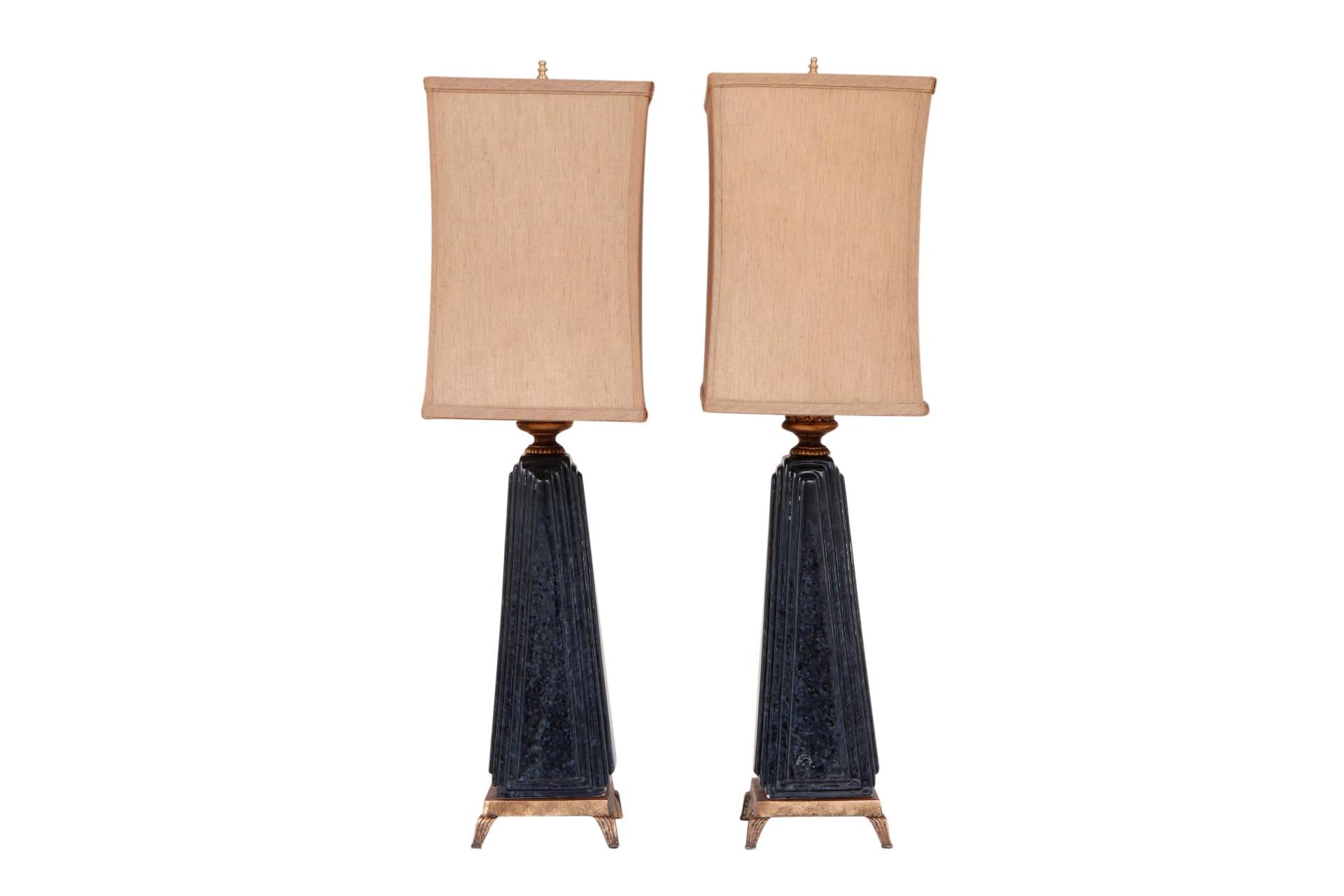 A pair of regency style ceramic table lamps. Lightly flecked navy columns taper upward and are squared with beveled edges. Sockets are decorated with outward flared acanthus leaves in brass. Square brass bases stand on reeded feet. Lamps come with