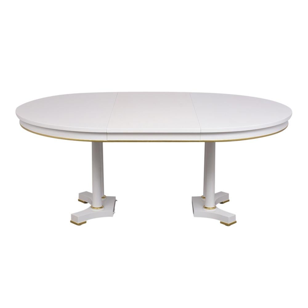 This 1970s Oval Dining Table by Baker furniture has been newly restored, is made out of solid mahogany wood, and has been newly stained a white color with a lacquered finish. The Dining Table has an extendable top with gilt accents along the edge,