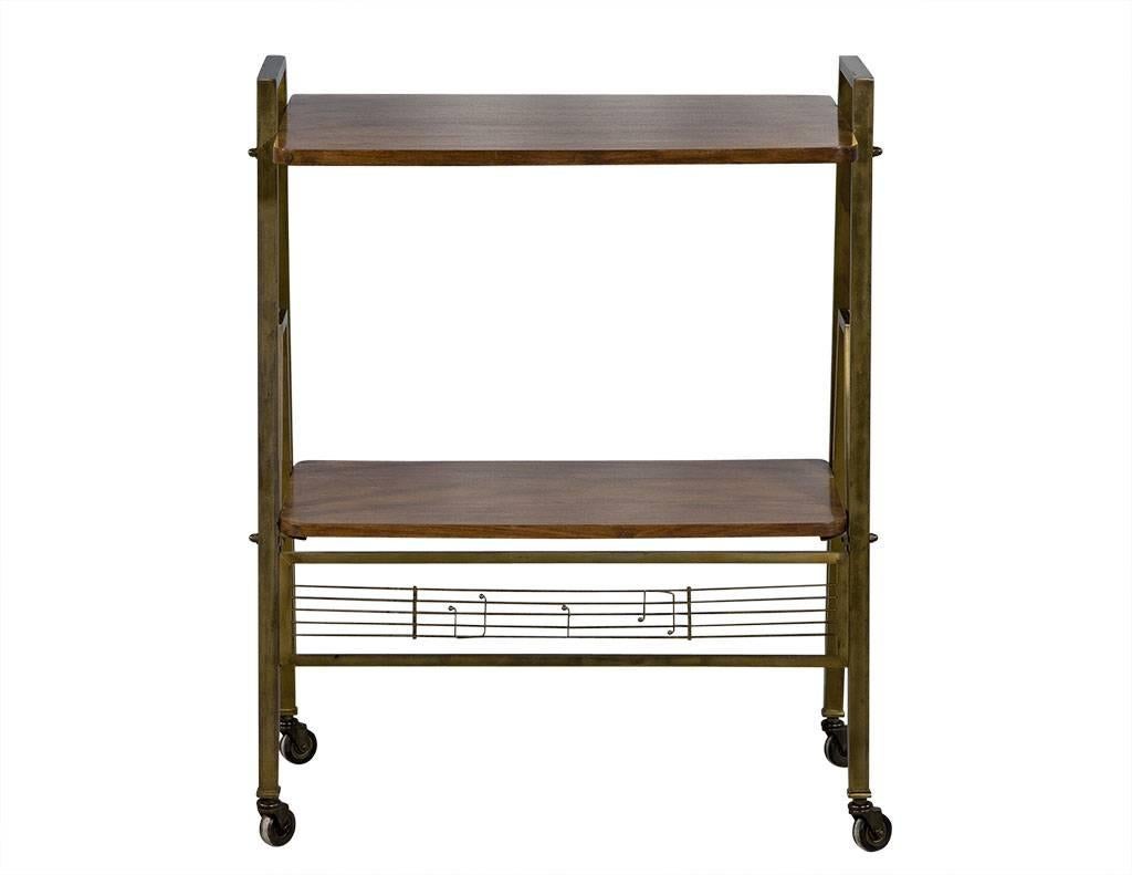This 1960s Regency style bar cart is in excellent condition. It has a musical theme, with 2 levels of reddish-brown wood and gold metal frame. The entire piece sits atop caster wheels, and the front panel at the bottom is a metal musical staff with