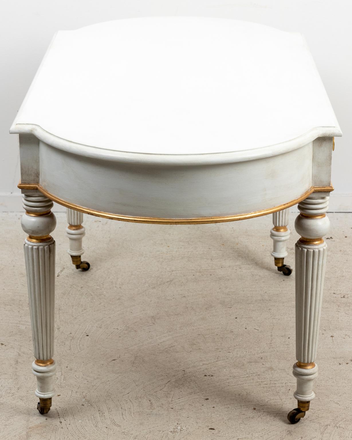 Circa 1980s Regency style white and gold writing table or robust desk with two drawers and working key. The piece also features curved ends with turned and reeded tapered legs ending on brass cup casters. Made in the United States. Please note of