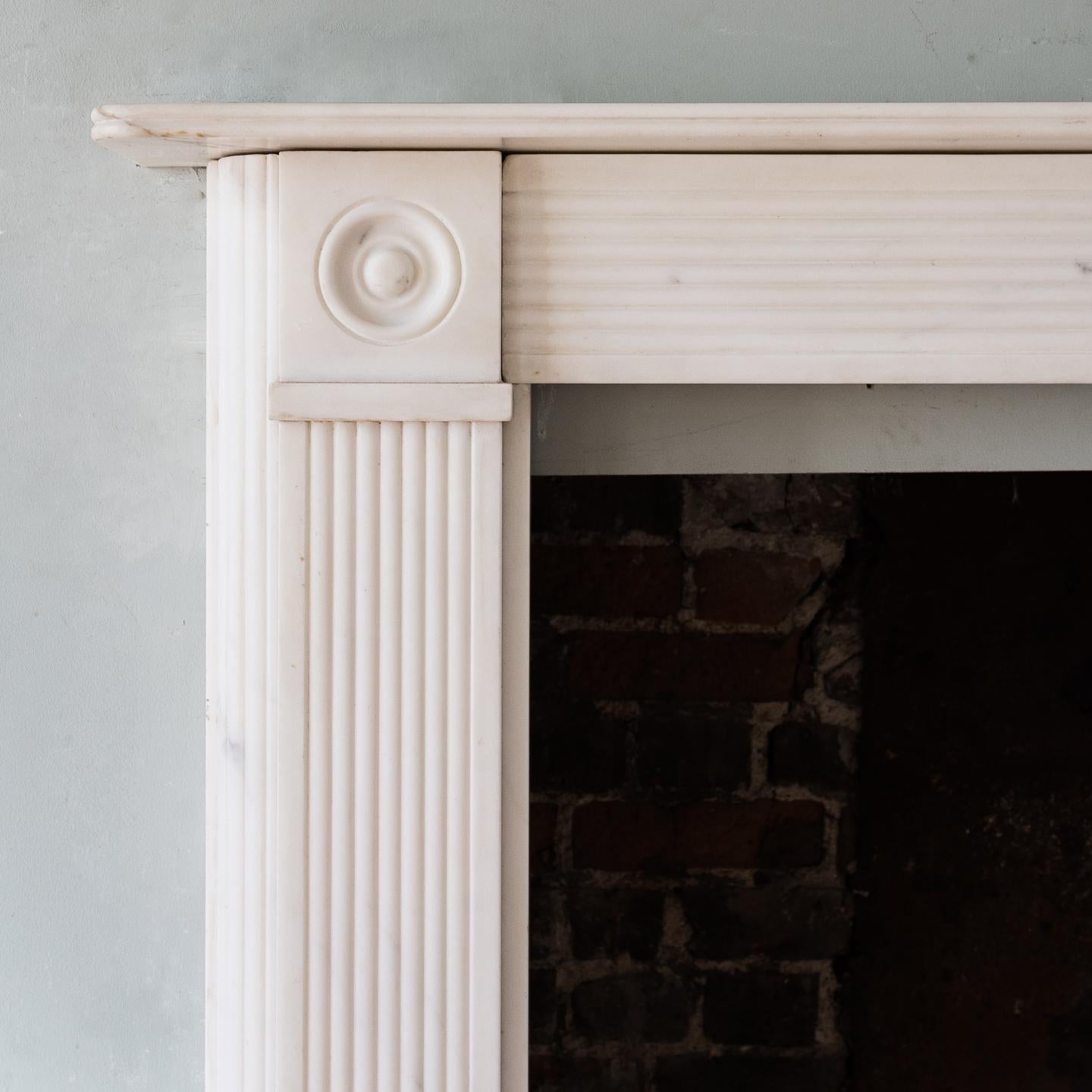 Regency style white marble bullseye chimneypiece, 20th century, the reeded mantle above reeded frieze centred by rectangular fluted plaque flanked by bullseye roundels leading to reeded jambs with additional curved reeded detail to the outsides,