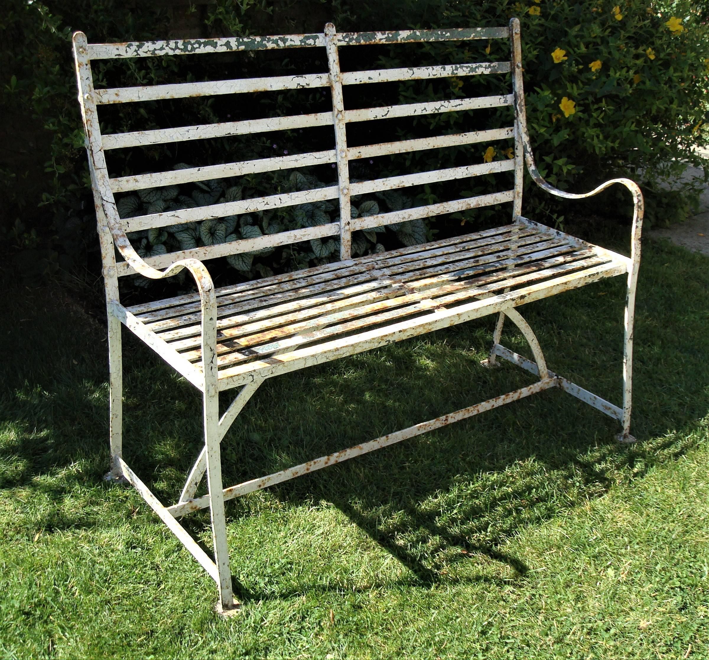 Good Regency wrought iron, slatted garden seat or bench of small proportions; the gently sloping back of slatted form with scroll down arms. The seat with close slats above a hooped stretcher, raised on a traditional 'H' stretcher; the legs