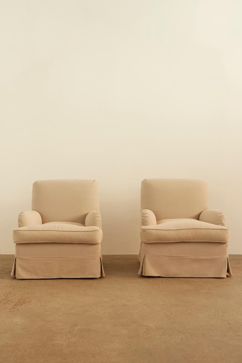 A pair of classic sand cotton slipcovered armchairs with a timeless silhouette. Featuring plush cushions and skirted bases, with beech feet, these chairs offer both comfort and elegance, making them a versatile addition to any interior.