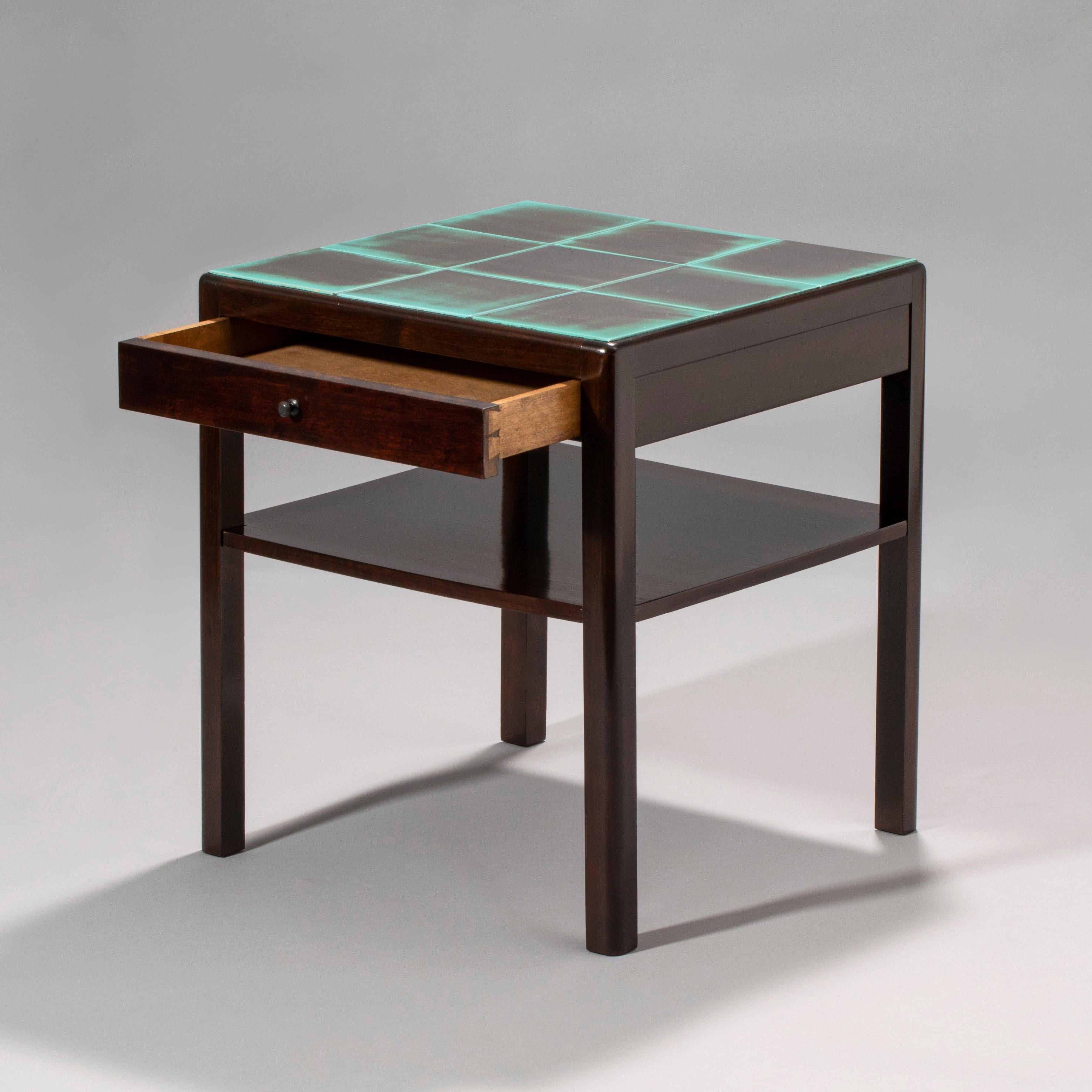 The square top composed of black and turquoise glazed ceramic tiles, set within a ebonized birch frame, the single drawer frieze centering a circular pull, above a shelf stretcher, raised on quarter-rounded legs. 

Stamped underneath with the