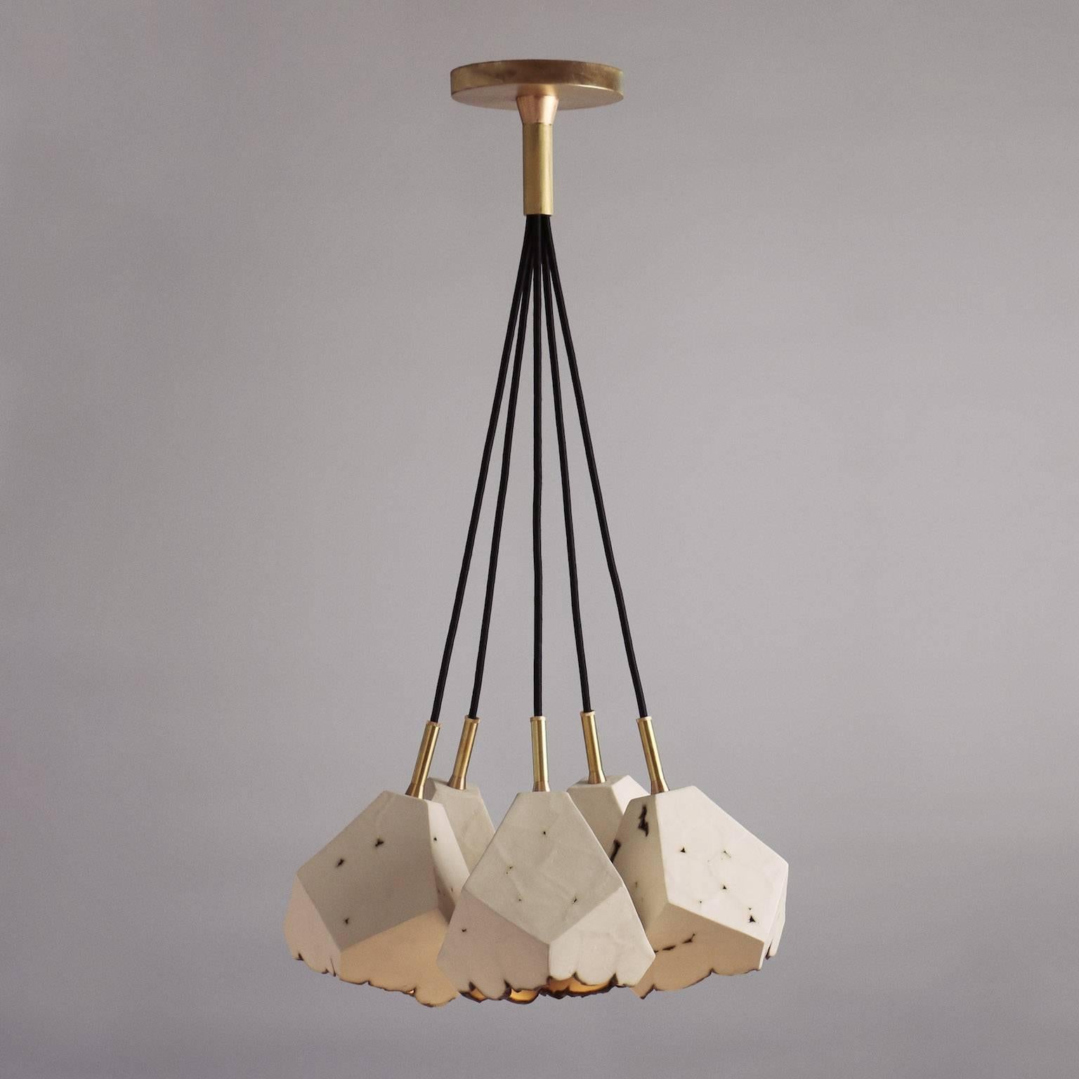 This sculptural chandelier features five geometric porcelain pendants with a unique textural matte finish, and custom brass hardware. The shades are each handcrafted from slabs of unglazed white porcelain with highly individual black oxide burnout