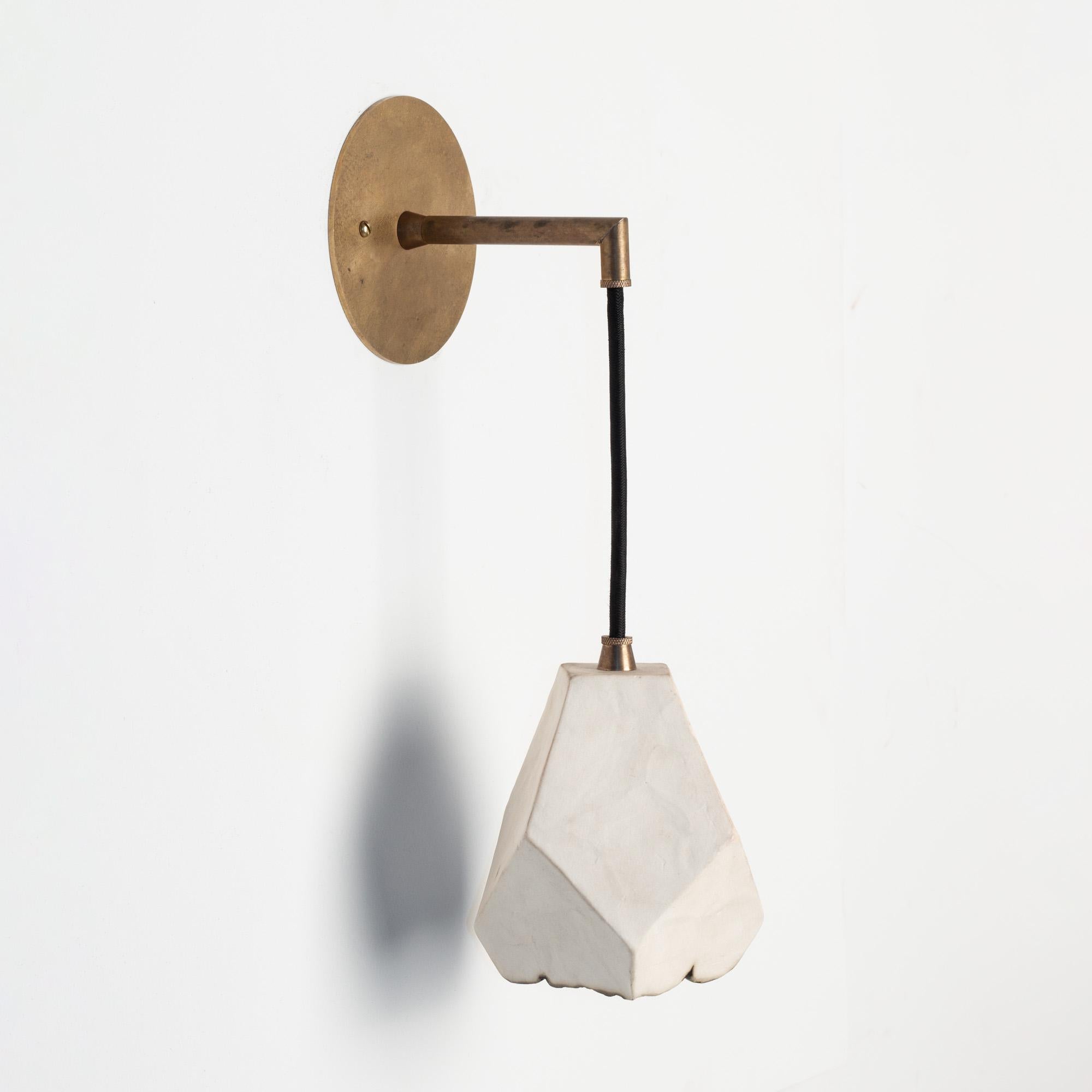 This sculptural wall sconce features a clean geometric shape and a unique textural matte finish. The shade is handcrafted from slabs of unglazed white porcelain with highly individual black oxide burnout detailing at the edges. The unfinished brass