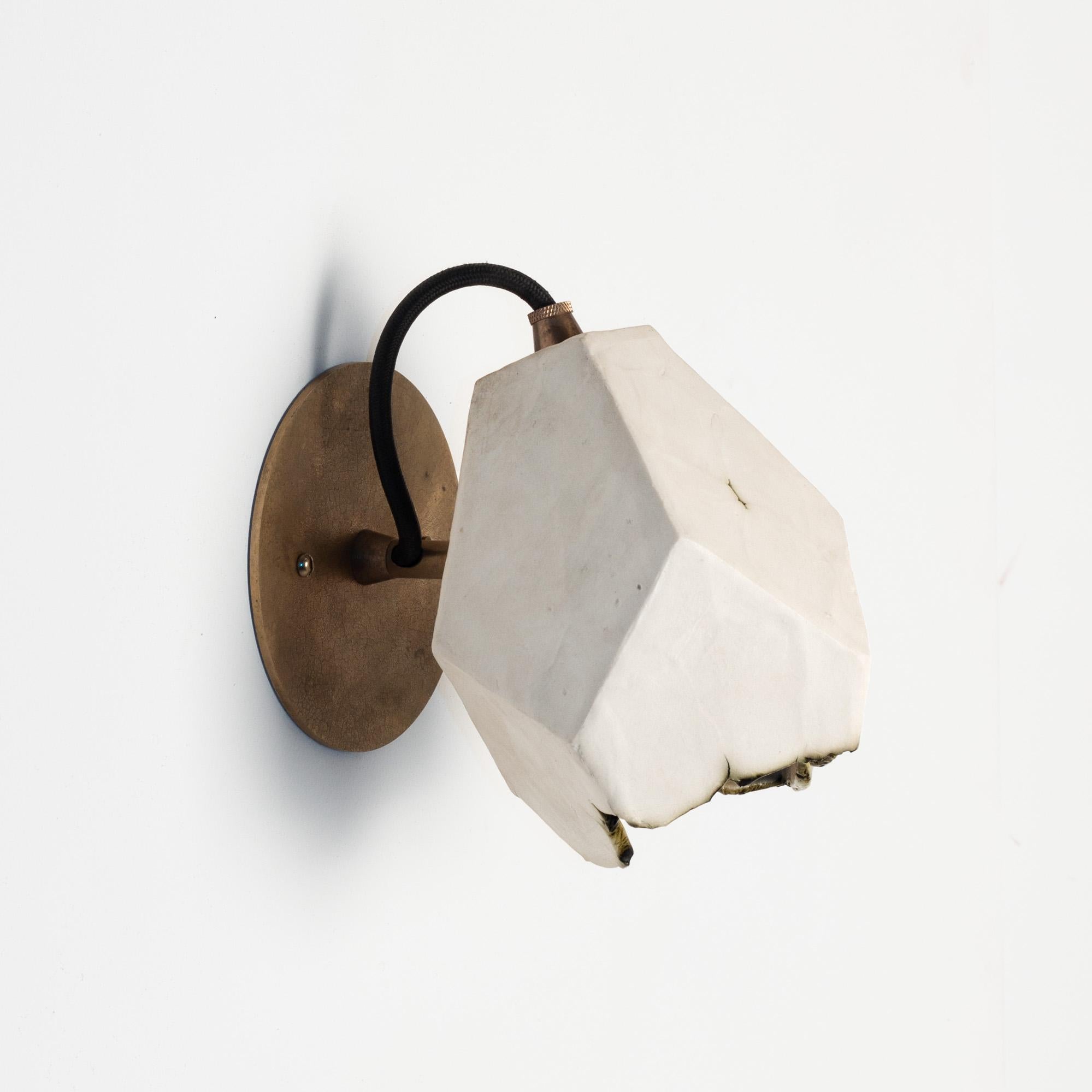 This sculptural wall sconce features a clean geometric shape and a unique textural matte finish. The shade is handcrafted from slabs of unglazed white porcelain with highly individual black oxide burnout detailing at the edges. The unfinished brass