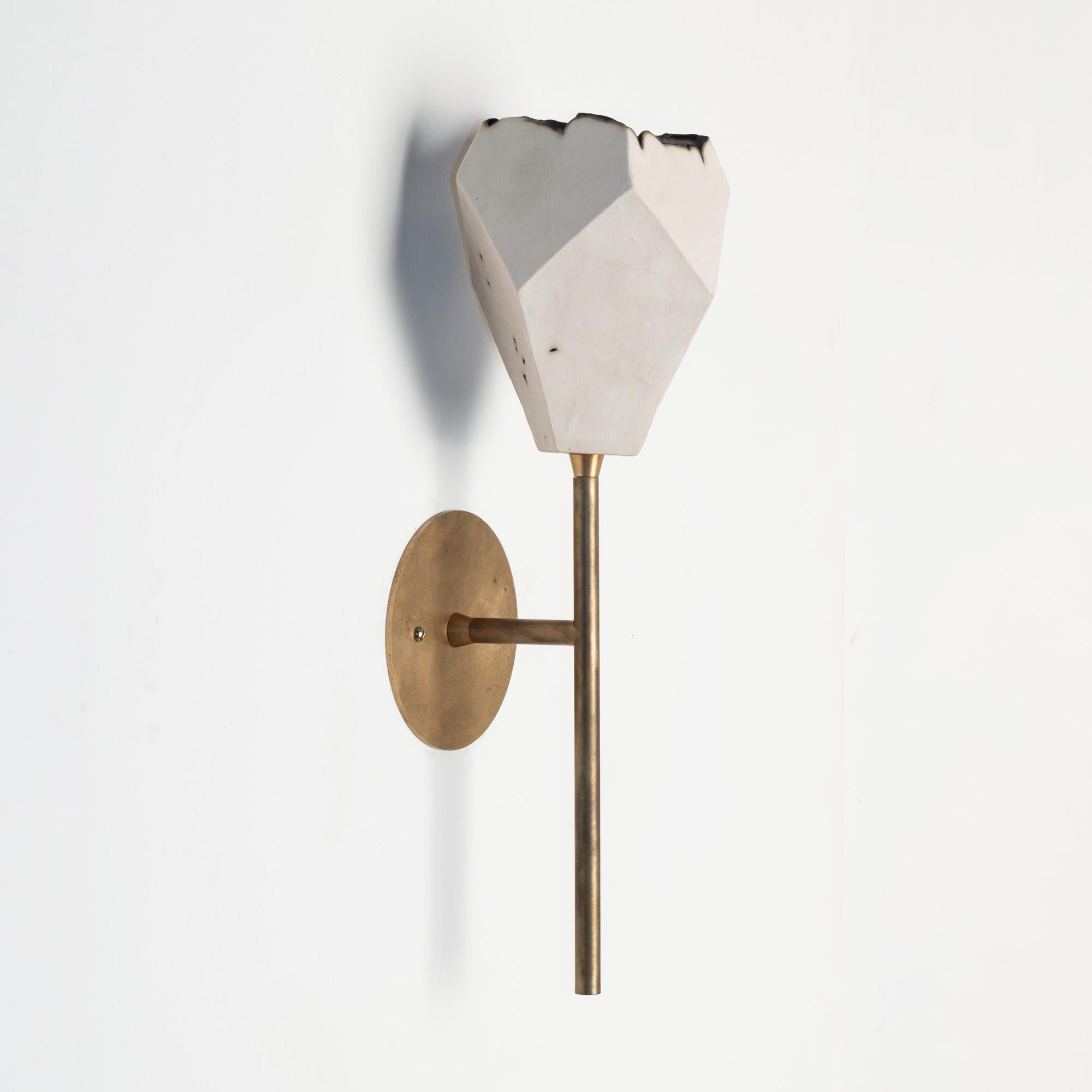 This sculptural wall sconce features a clean geometric shape and a unique textural matte finish. The shade is handcrafted from slabs of unglazed white porcelain with highly individual black oxide burnout detailing at the edges. Unfinished brass