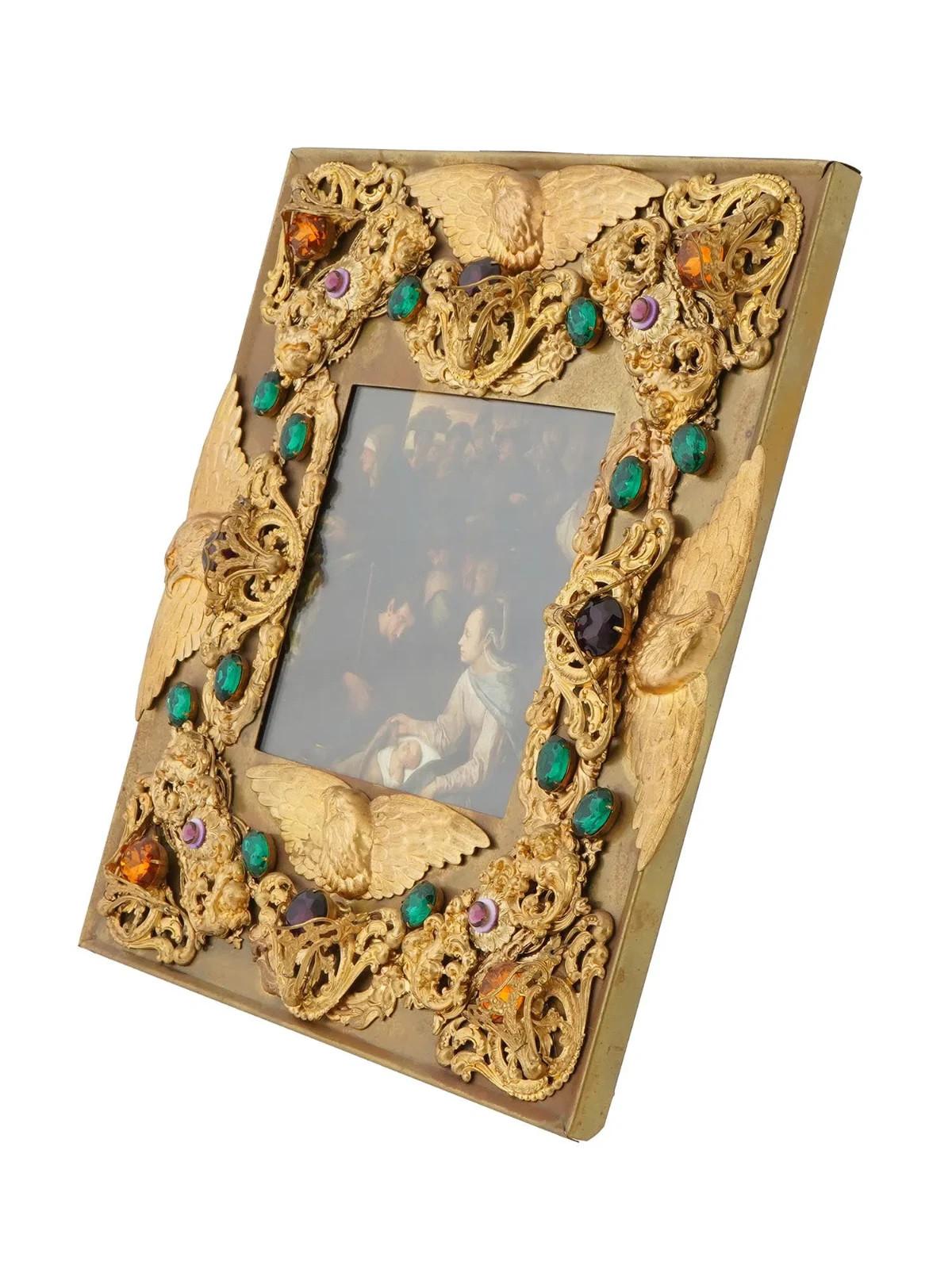 Vintage-Bilderrahmen aus vergoldetem Messing. Der Rahmen weist ein Reliefdesign auf, das mit dekorativen Elementen wie bunten Steinen auf einem goldfarbenen Hintergrund verziert ist. Das Geschirr ist mit geprägtem, verschnörkeltem Blattwerk und
