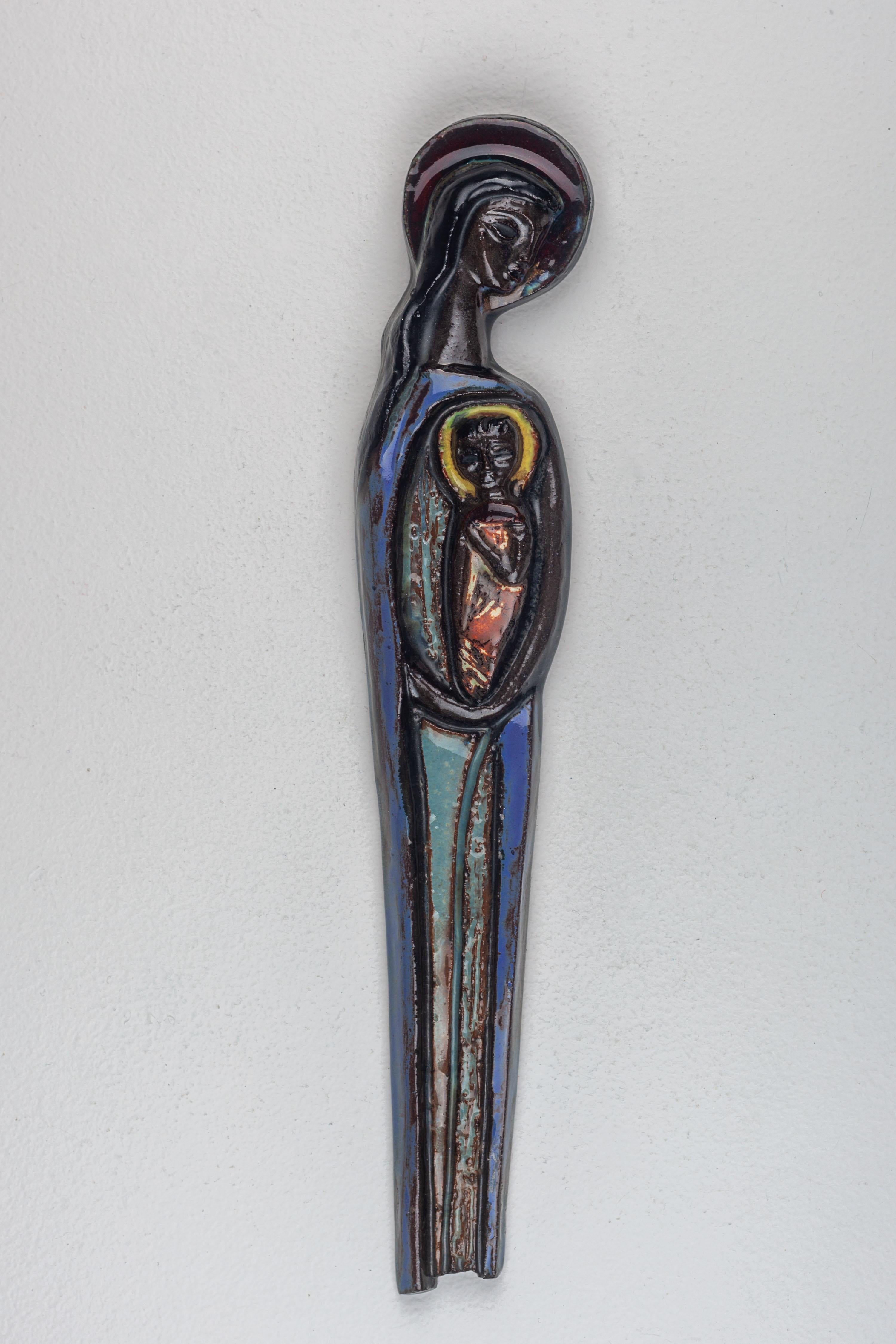 This piece of mid-century ceramic wall art captures the Virgin Mary as she cradles the Christ child. The sculpture is defined by its deep, resonant color palette, utilizing rich blues and stark blacks finished with a semi-gloss glaze that subtly