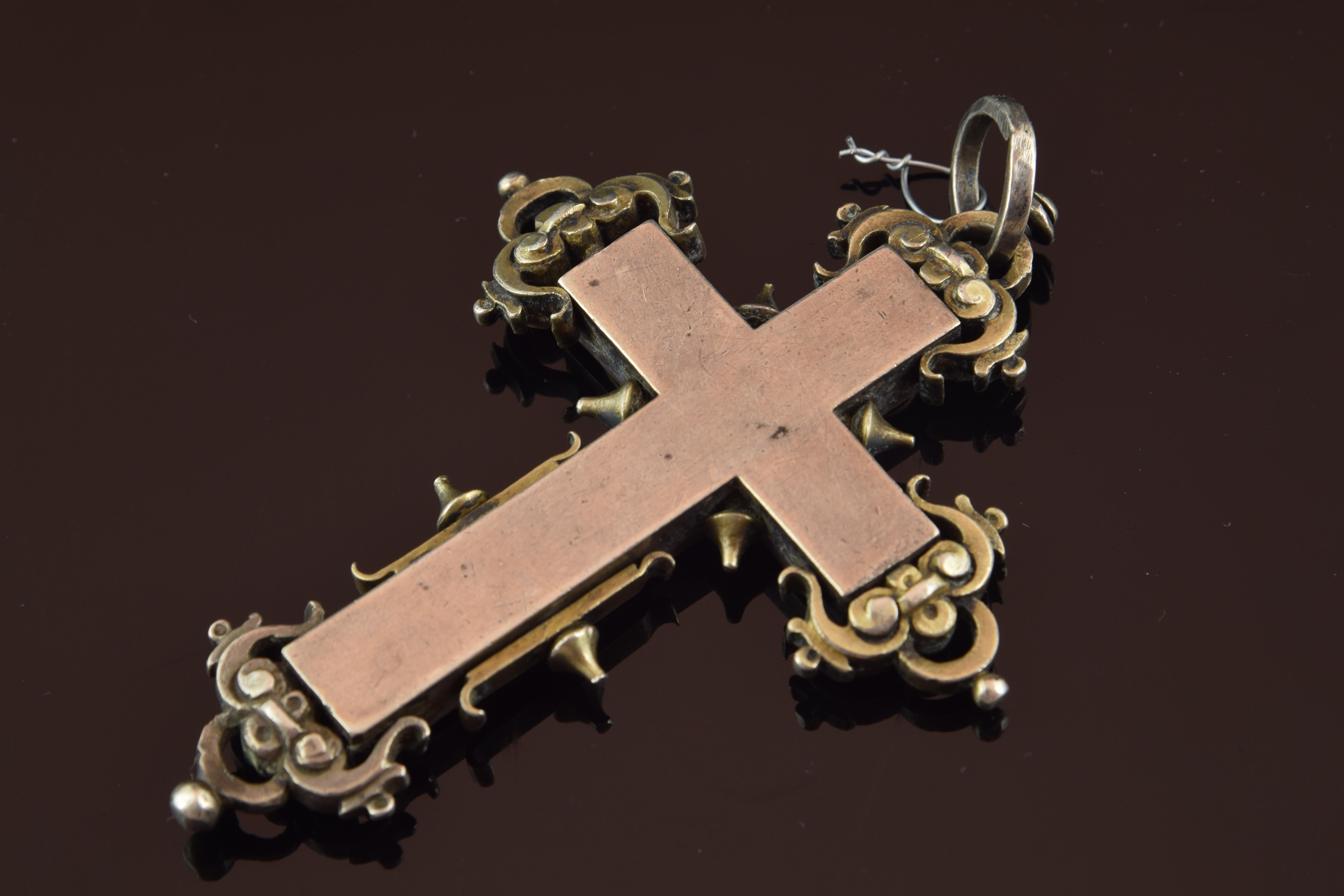 Devotional pendant made in silver with a Latin cross shape, decorated on the outside with simple scrolls, spikes, pearls and other elements of classicist inspiration, as well as a ring to wear the jewel hanging from the neck. The front has seven