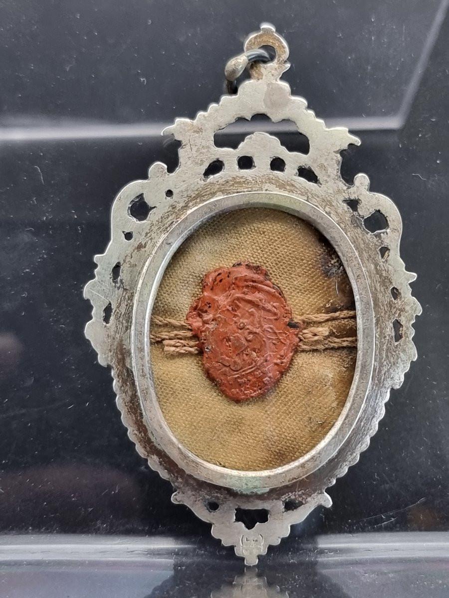 Pendente-reliquiario in argento fuso e cesellato, di gusto barocco-rocaille, con cornice traforata e vetro frontale. Sul retro è presente un vetro trasparente con sigillo in ceralacca rossa attraversato da cordoncini originali, ancora integro.