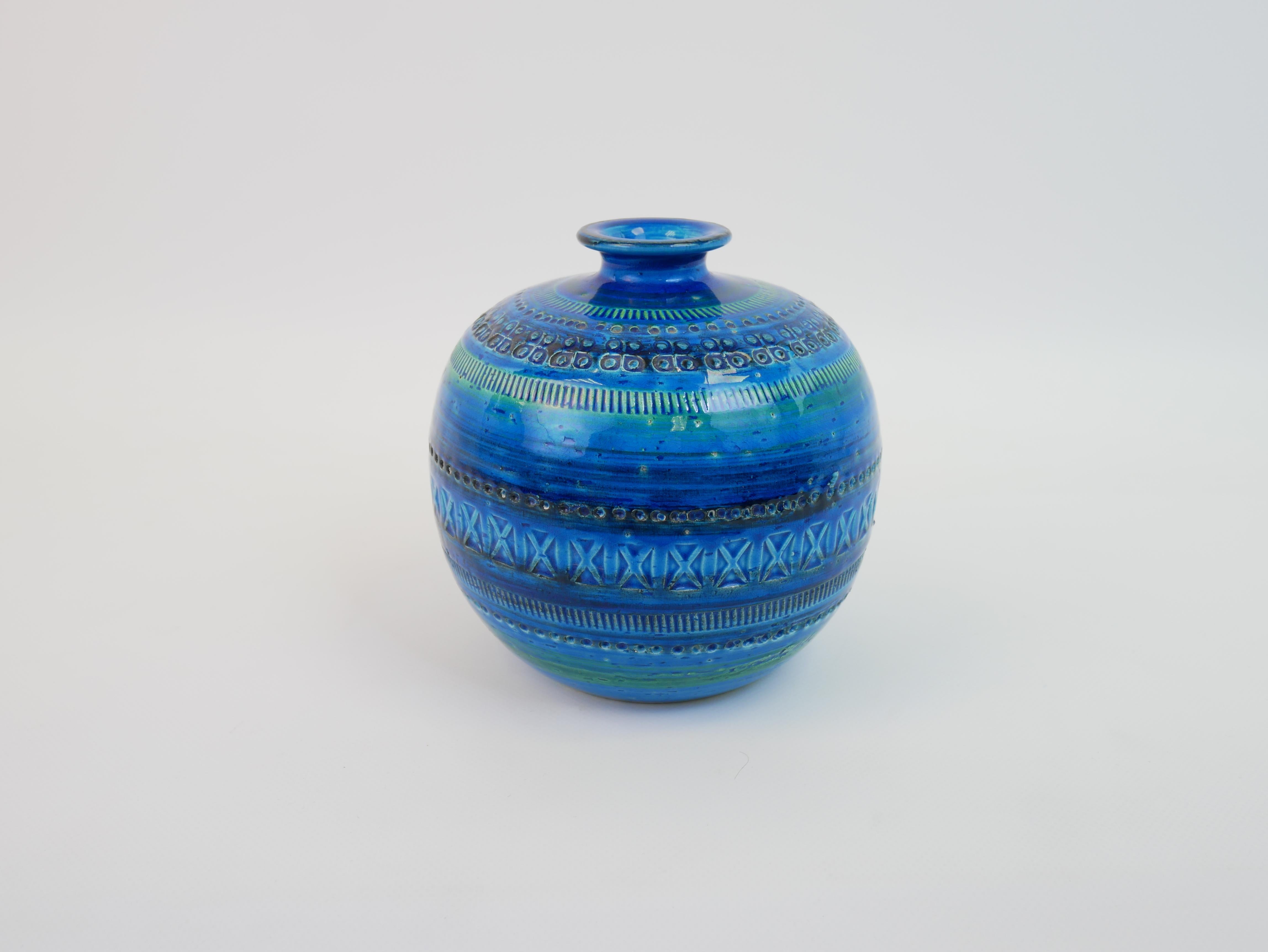 This Flavia Italy ceramic vase features a round shape with a narrow neck. The glaze is a striking mix of deep blue and turquoise, accented with subtle green shades. The surface is richly textured with intricate geometric and abstract patterns,