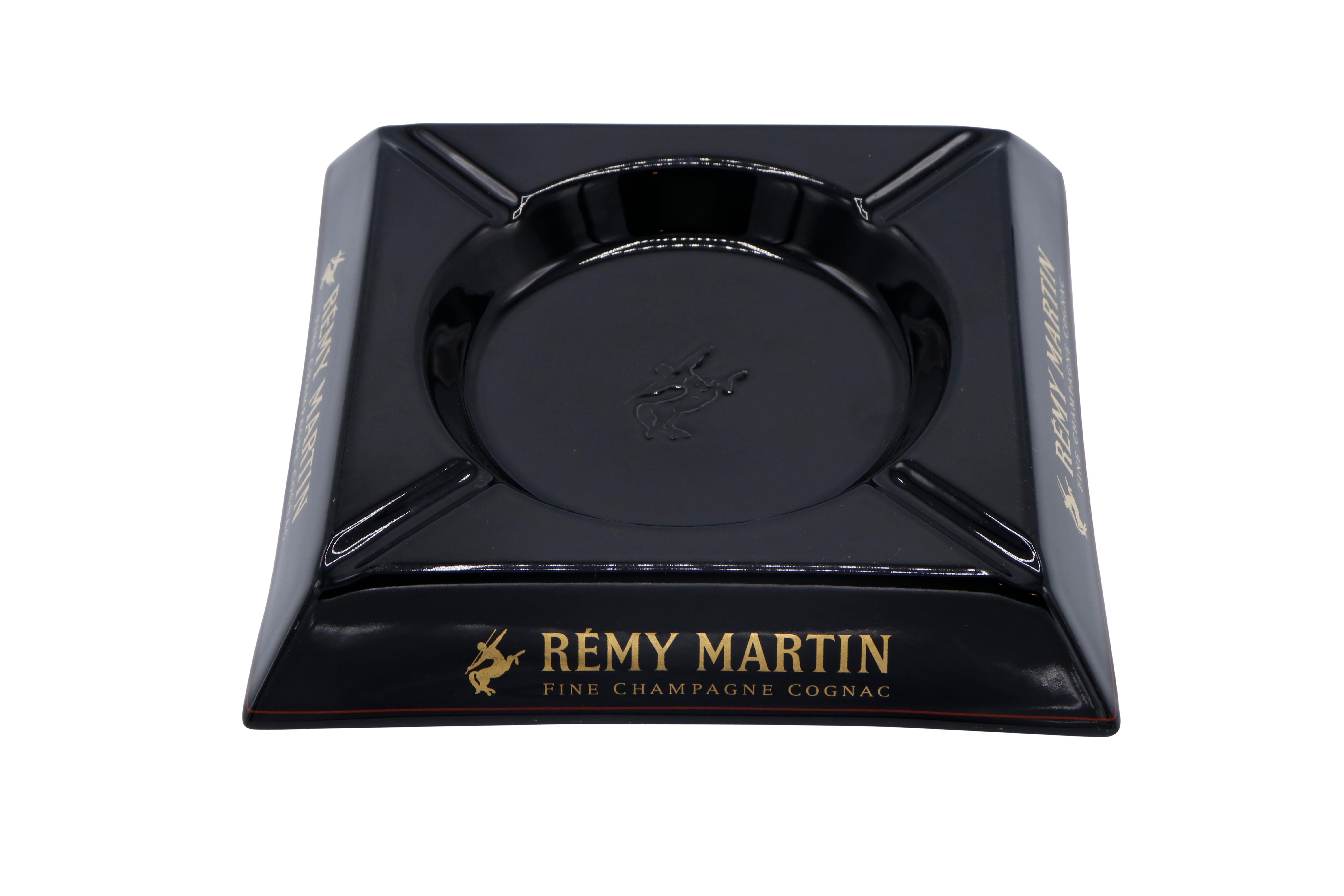 A French black square ceramic ashtray made by Rémy Martin. A central circular tray is embossed with the Rémy Martin logo, a centaur wielding a spear. Each corner has a beveled cigarette rest and the sides are decorated with the brand name and logo