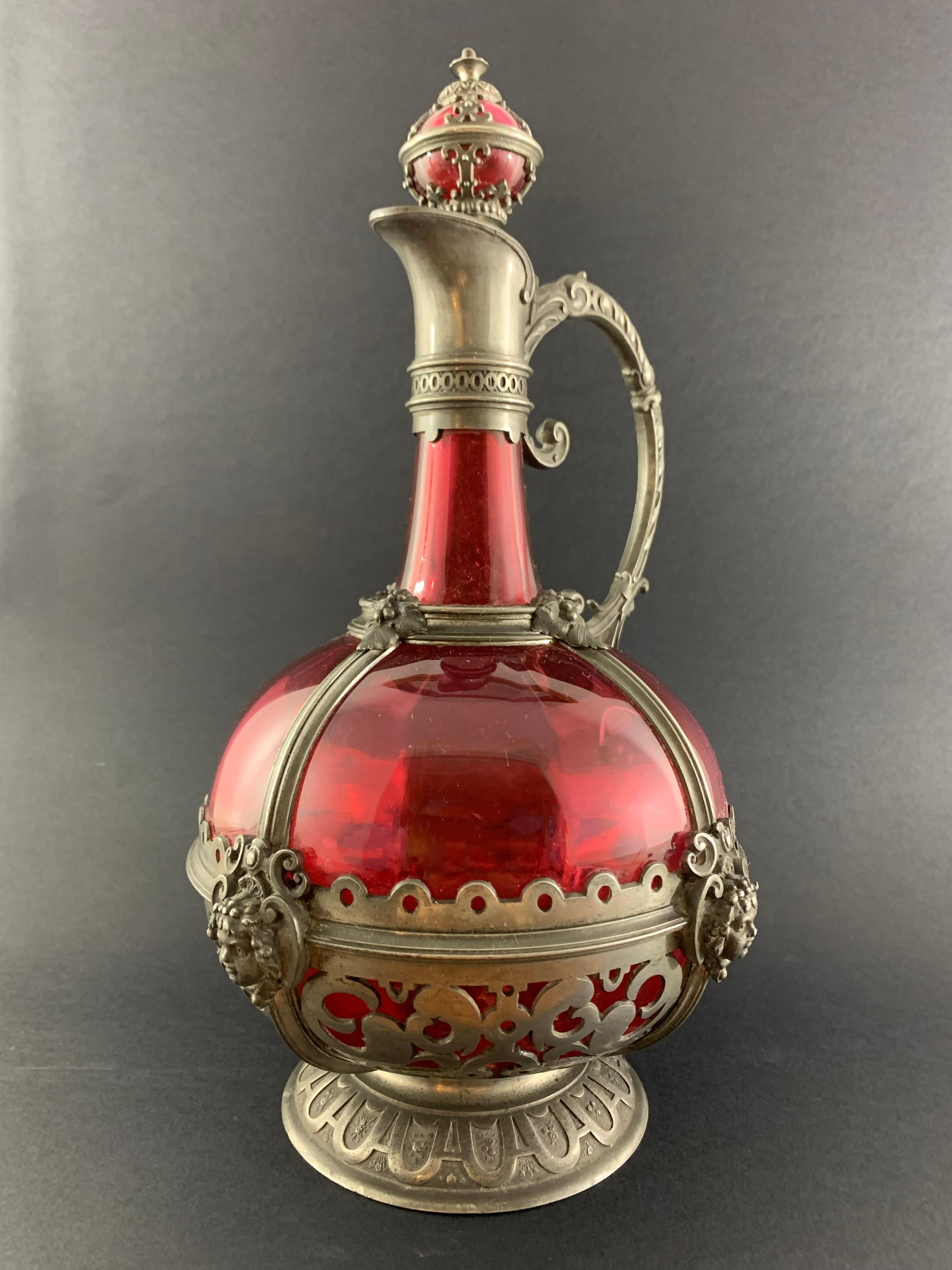 Beautiful red crystal ewer with pewter frame. We find on it motifs of vine and grape recalling a banquet, the drink. Four faces on each side are directly linked to Dionysus, the god of the vine, wine, excess, madness and excessiveness.
The 3.93