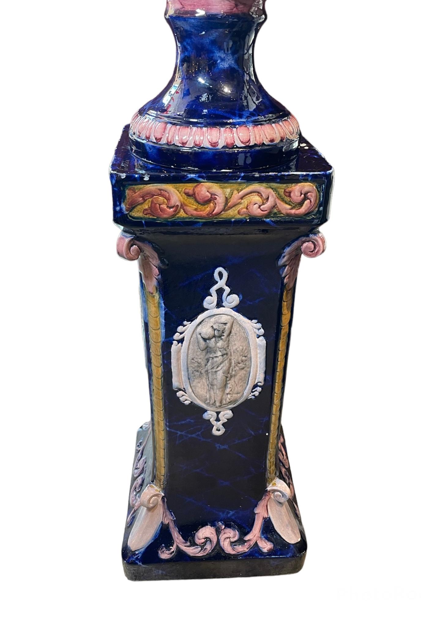 This is a Renaissance revival style large Majolica amphora vase with rectangular pedestal. Their background are hand painted cobalt blue and decorated with an off-white relief of Greek/Roman scenes. The amphora is adorned with salmon color scrolls