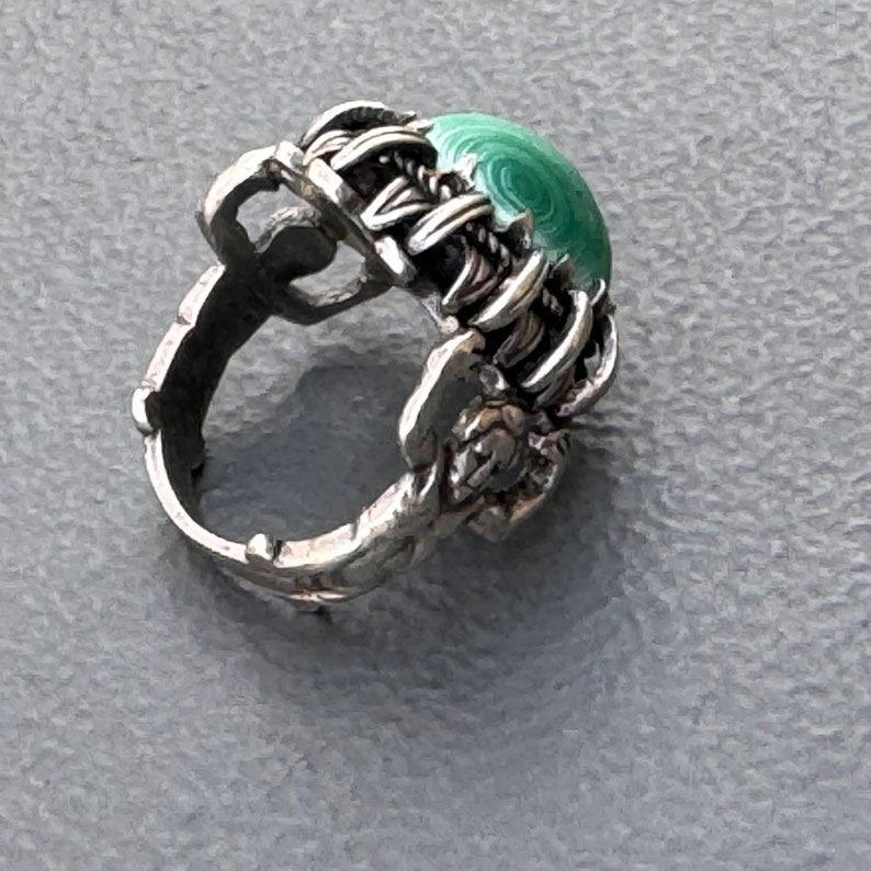 This captivating ring, dating from the late 19th to early 20th century, epitomizes the Renaissance Revival style with its intricate craftsmanship and dramatic design. The ring features a striking malachite stone set in an ornate silver setting,