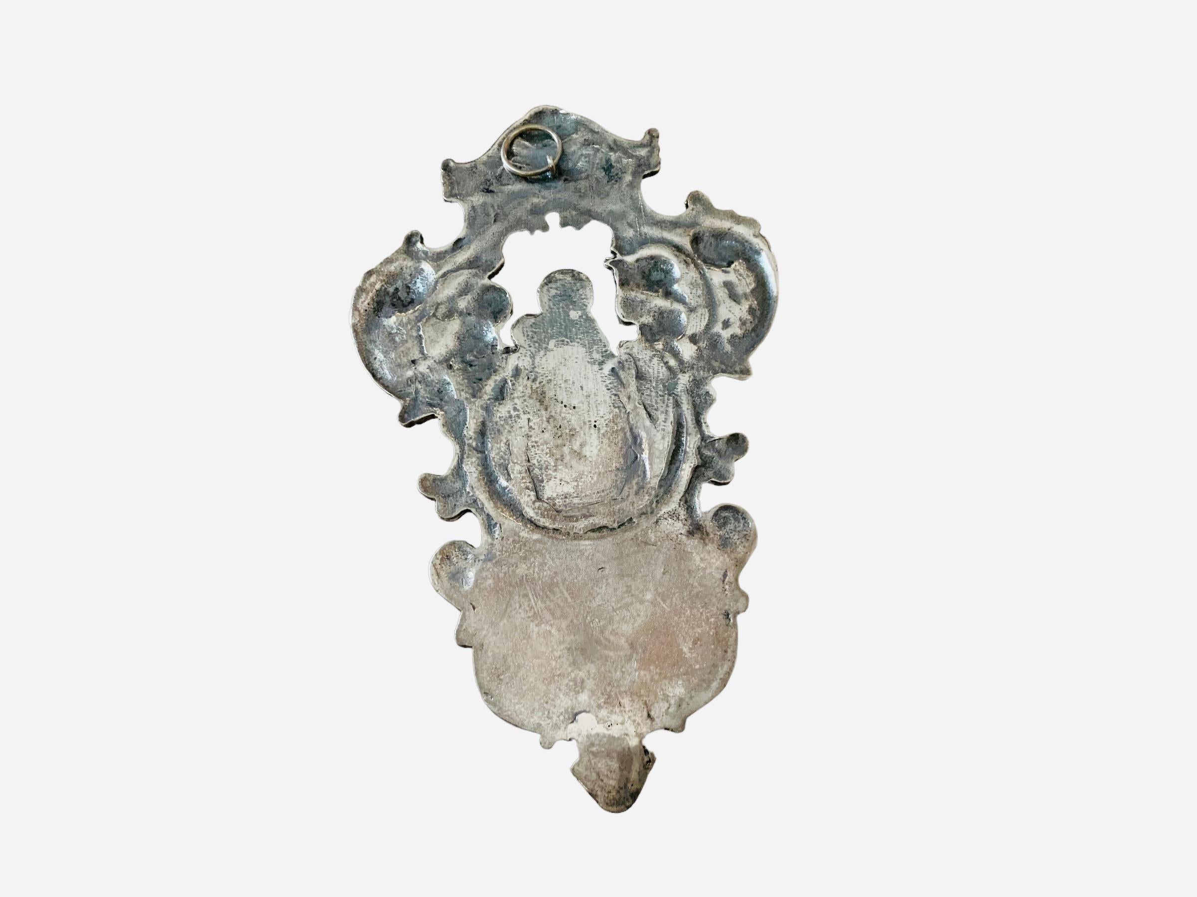 This is a small silver benitier. It depicts the relief of Virgin Mary holding Baby Jesus in the upper center of the benitier that is adorned with scrolls of acanthus leaves and flowers around them. An embossed pairs of cherubs are each side of them.