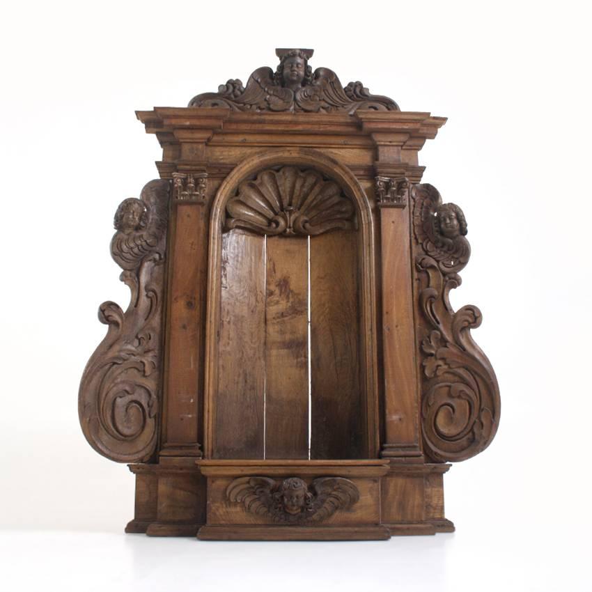 Symmetrically designed wall nook with a rich carved décor in the shape of scrolls, shells and winged angels. Pilasters with Corinthian capitals flank the semi-circular nook.