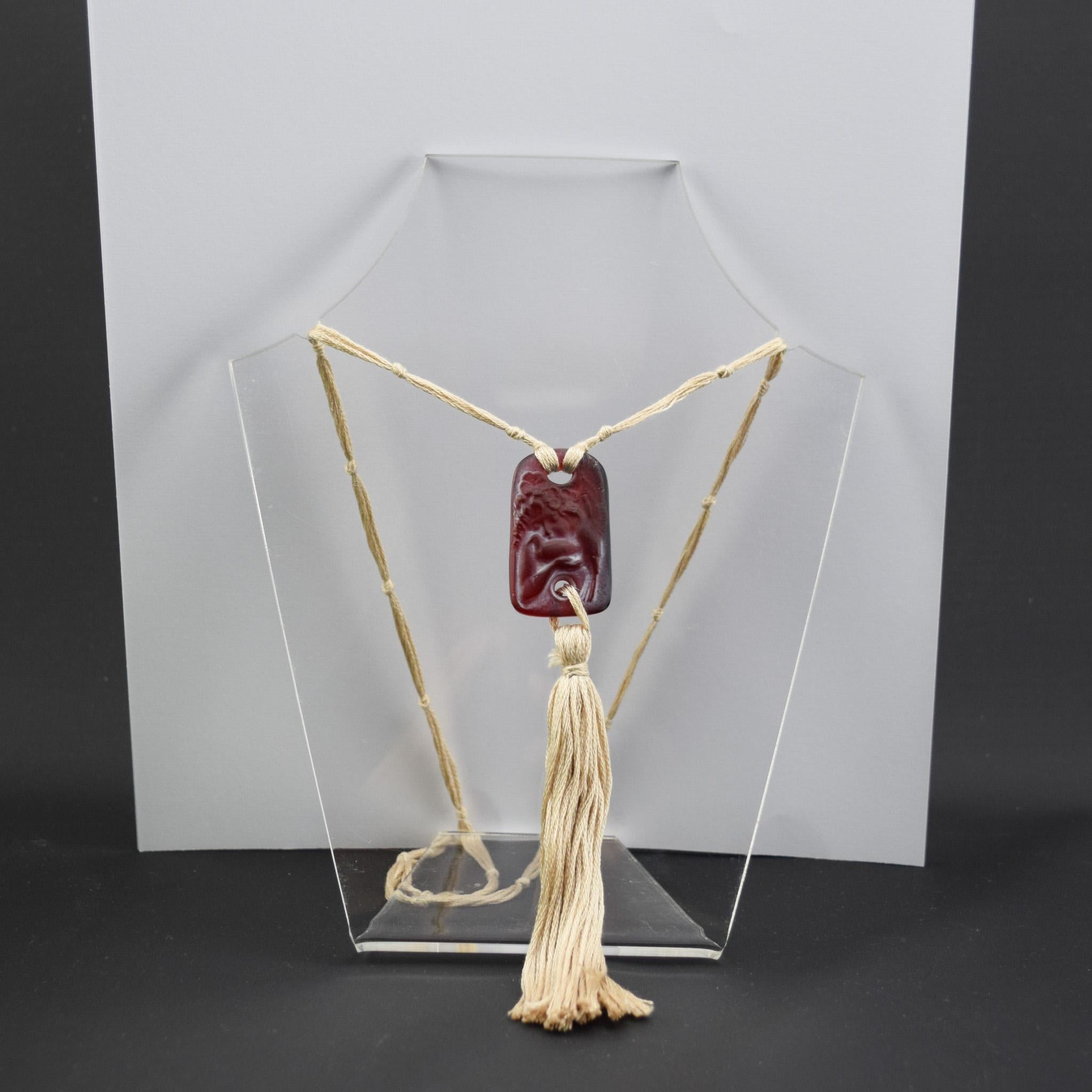 Colombes-Anhänger von Rene Lalique aus rotem Glas mit weißer Färbung, der ein Kind mit zwei Tauben darstellt

Aufgereiht mit beigefarbener, geknoteter Seidenkordel und Quaste

Signiert R Lalique auf der Rückseite

Marcilhac Seitenangabe 580
Modell