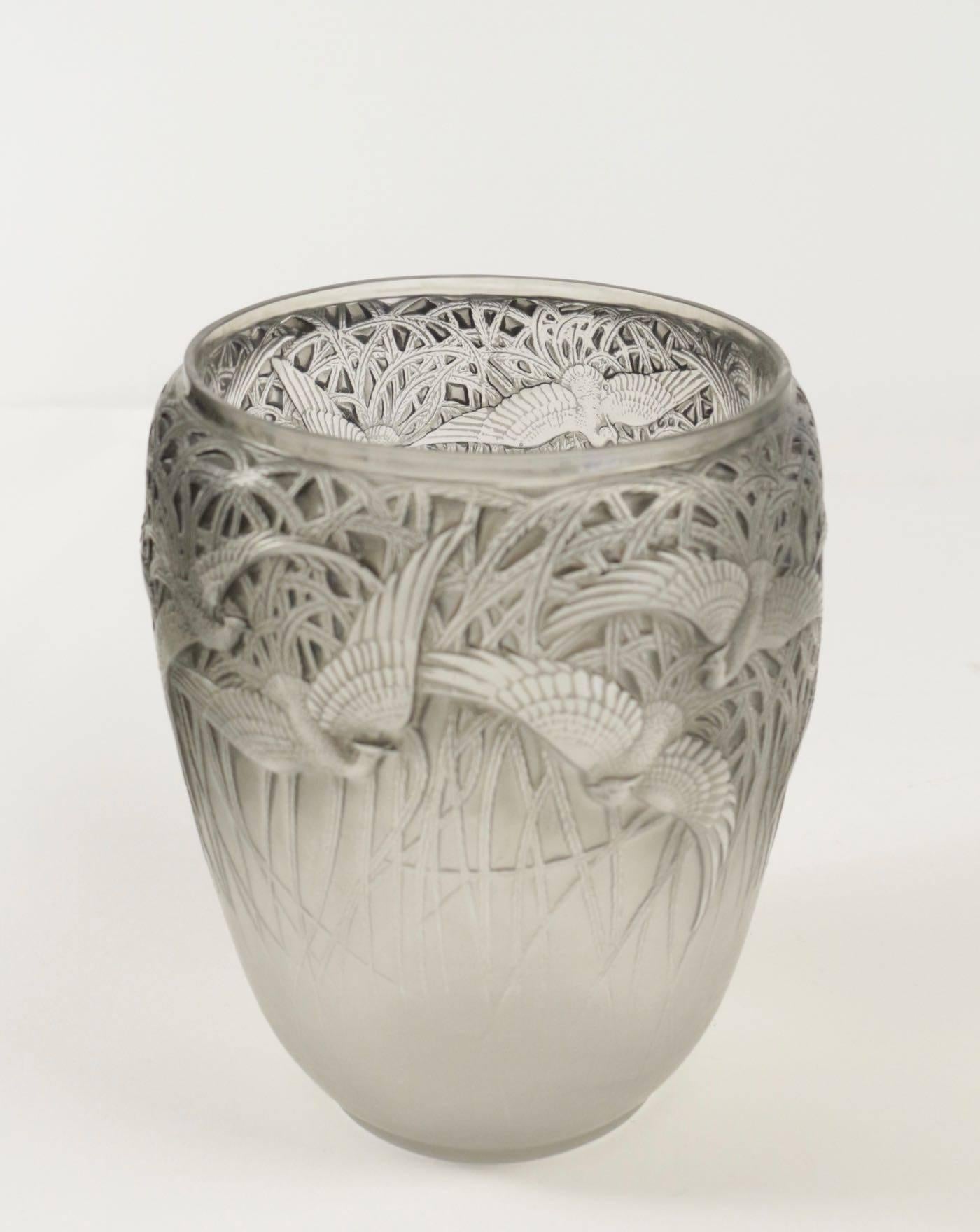 René Lalique (1860-1945)
vase Aigrettes.
Measures: 8 and 1/2 inches high.
Mold pressed glass having a flying egrets against a tall reed style plants background all around the exterior.
Glass highlighted with blue grey patinated.

Model created