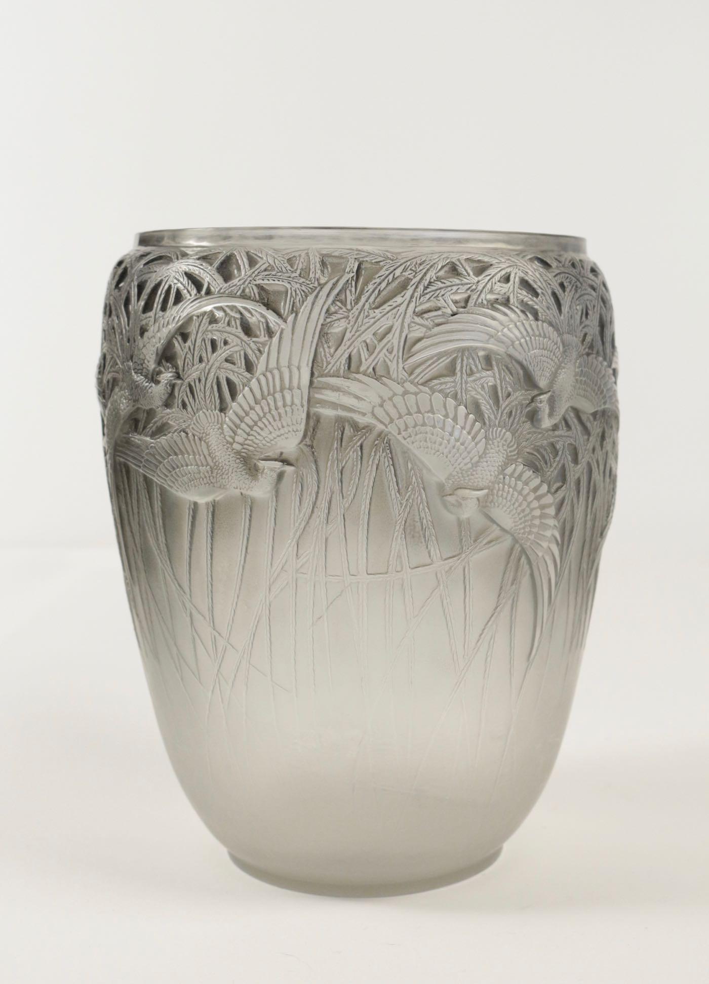René Lalique (1860-1945)
Vase Aigrettes.
Measures: 8 and 1/2 inches high.
Mold pressed glass having a flying egrets against a tall reed style plants background all around the exterior.
Glass highlighted with blue grey patinated.

Model created
