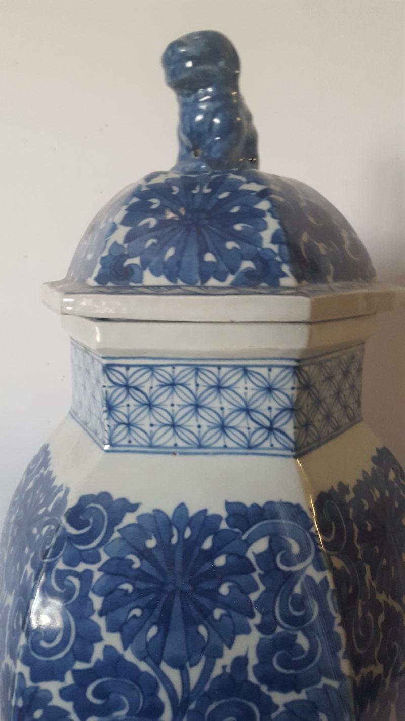 This large, decorative, faceted jar or container is made of white glazed pottery, covered in a traditional, Chinese blue floral design. It has a removable, glazed lid in the same applied design and topped by a foo dog finial or handle in solid blue