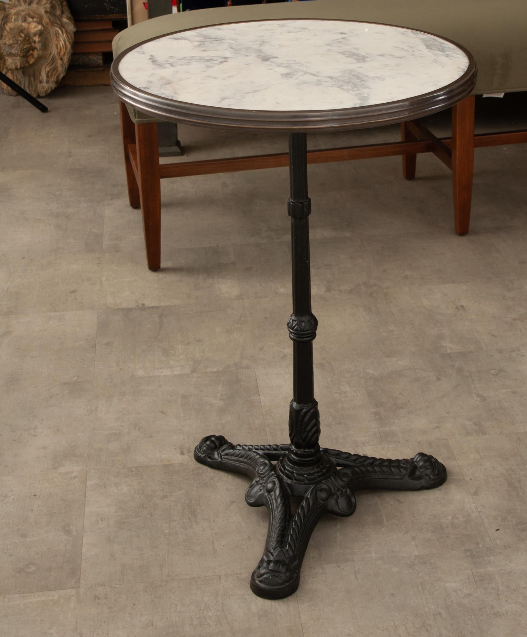 Ein klassischer Bistrotisch im Pariser Stil mit Marmorplatte und Eisen. Die einzigartige Platte aus weißem Marmor wird von einer Zierleiste aus antikem Messing umrahmt. Die Basis ist ein klassischer gusseiserner Sockel mit drei Füßen. Dieser Tisch