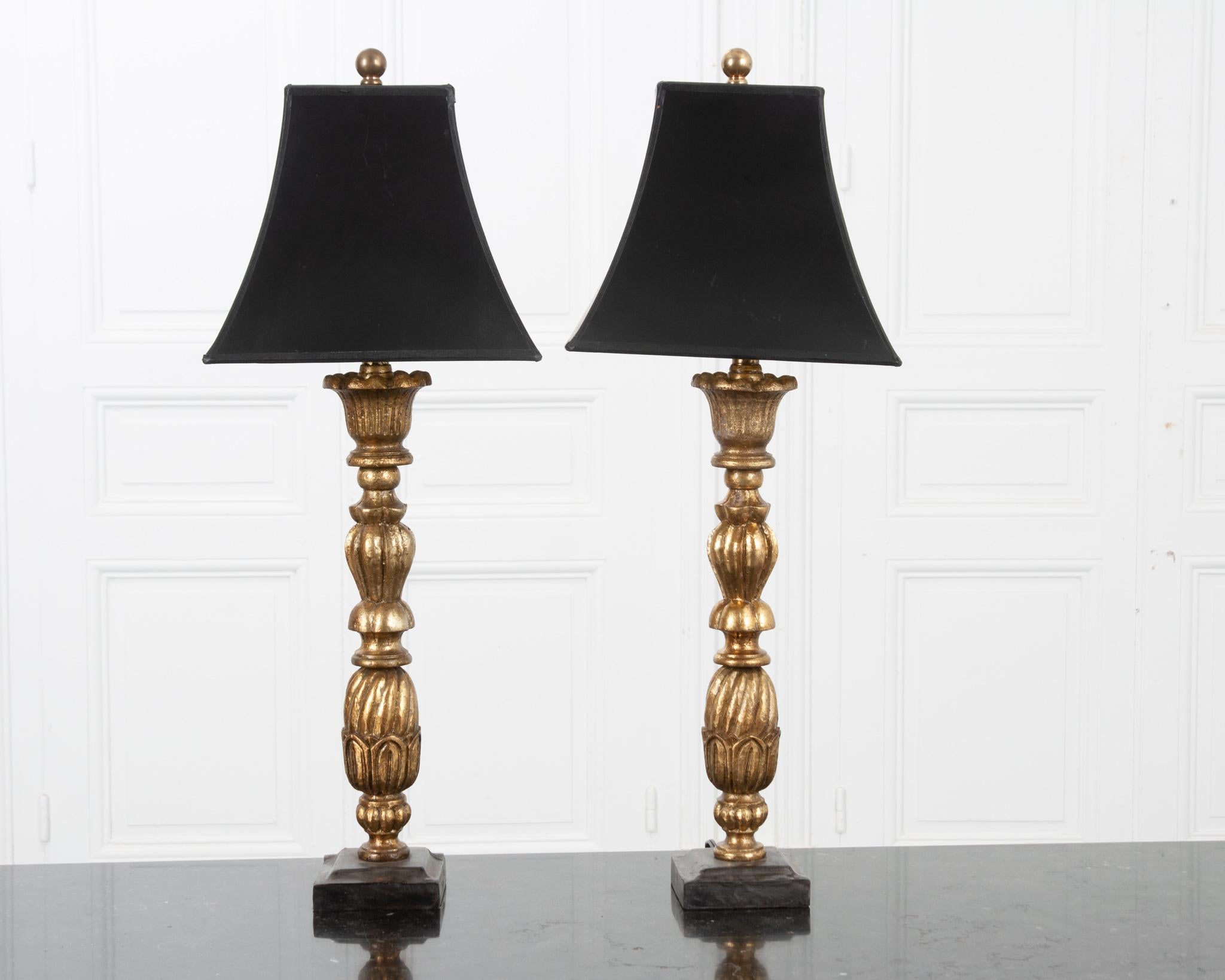 These hand carved library lamps are fixed with black paper shades that beautifully contrast the lustrous gold gilt bases. They feature deeply carved acorns and organic shapes on painted metal bases. Circular brass finials secure the shades. The