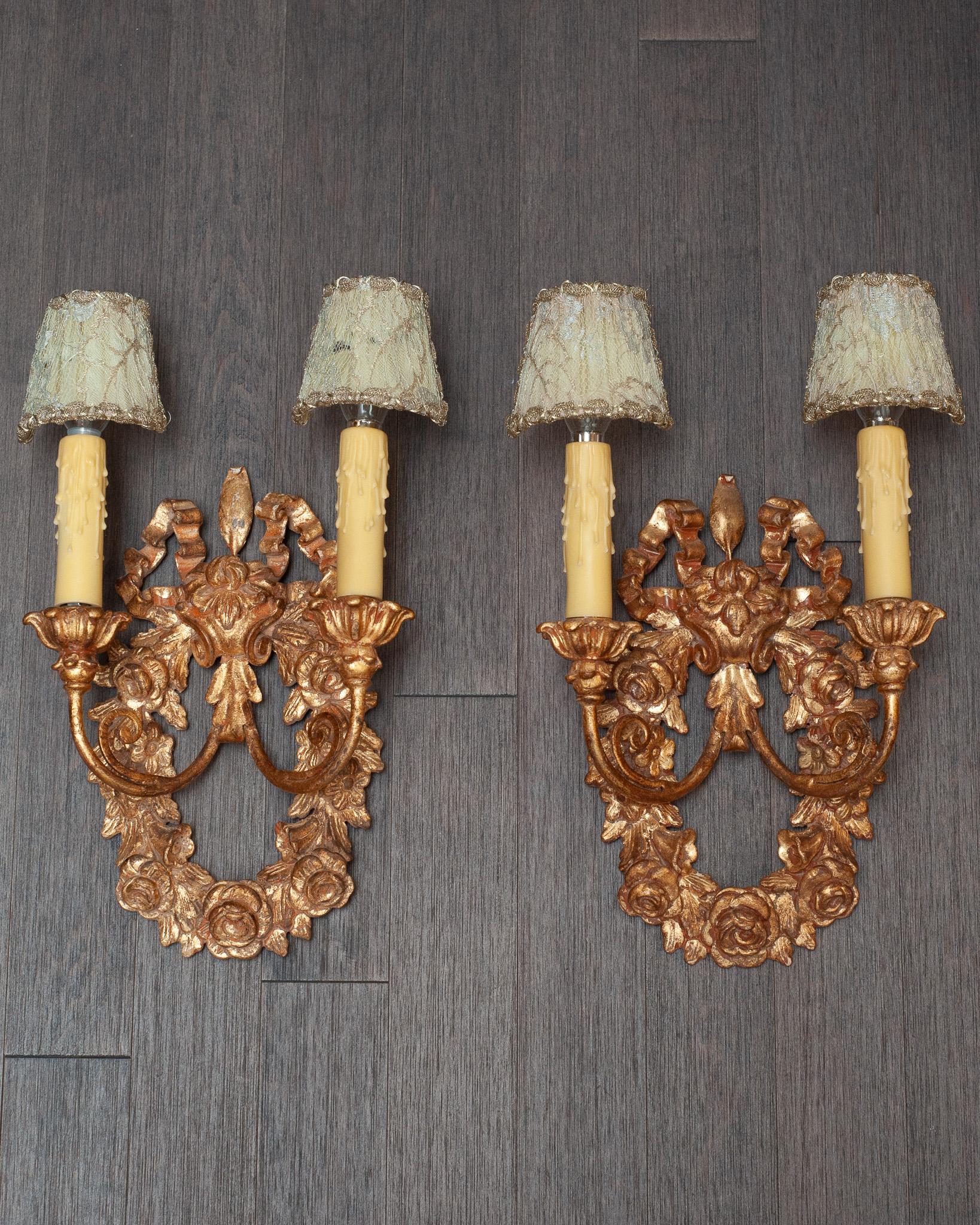 A beautiful pair of reproduction giltwood wreath sconces, with lace overlay silk shades and metallic trim. Each sconce wreath is filled with roses and flowers, with a ribbon motif on the top. The wooden back of each sconce measures 7
