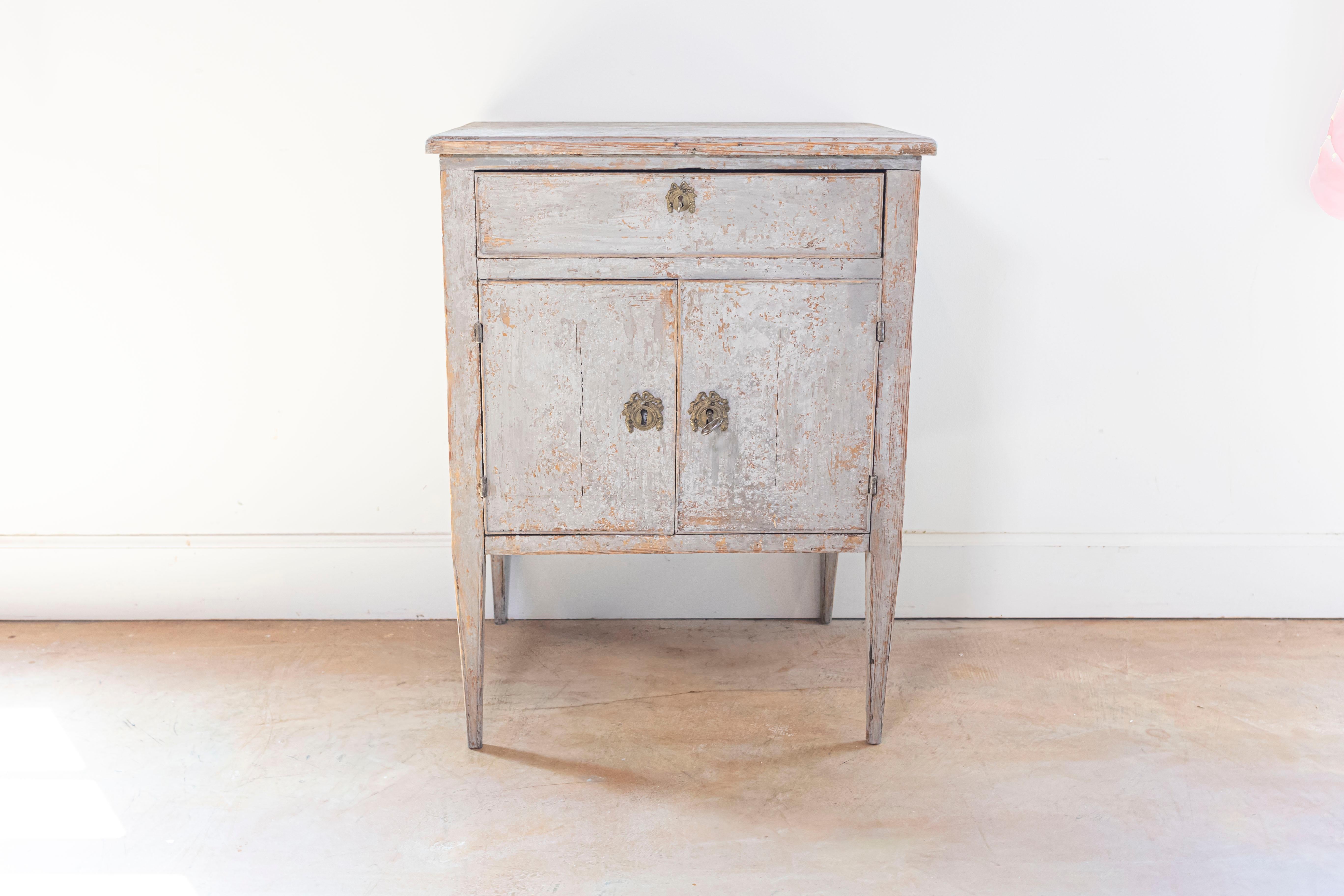 A Swedish Gustavian style painted bedside table from the 19th century with single drawer, double doors, tapered legs and ornate hardware. Immerse yourself in the timeless elegance of the 19th century with this Swedish Gustavian style painted bedside