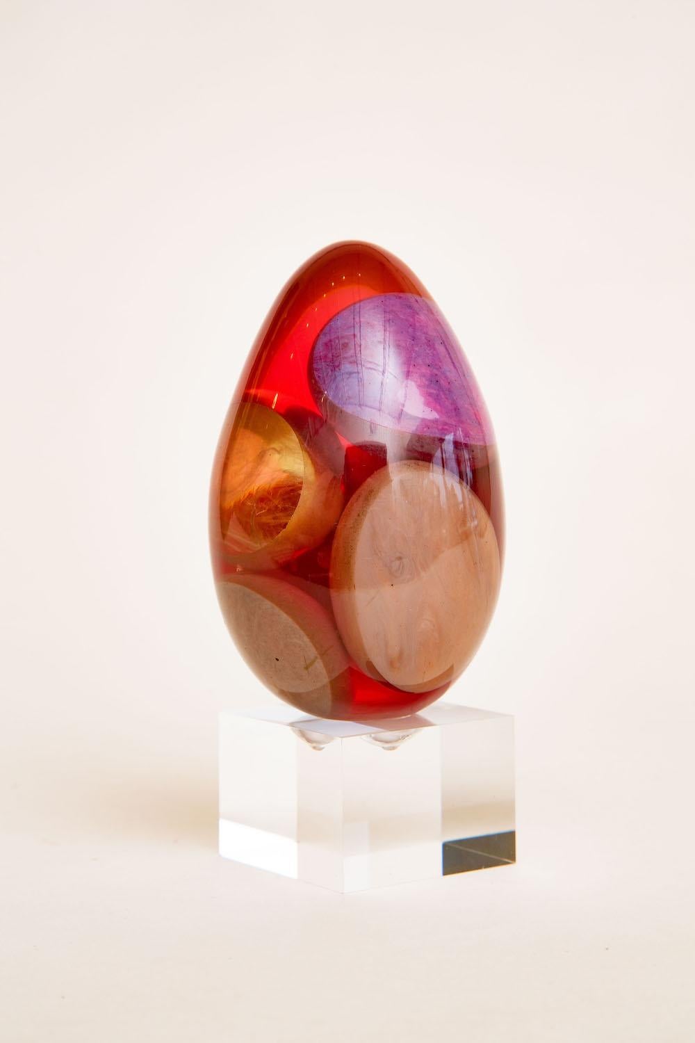This wonderful unusual vintage small resin ovoid egg shaped sculpture has floating resin pieces inside in large shapes of the colors of purple, tan, gray, yellow and translucent inside the red resin. The red casts a color of magenta red. Each side