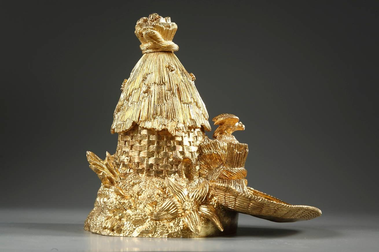 Small, gilt bronze inkwell in the shape of a hive. The inkwell cover is decorated with sheaves of wheat with bees resting on top. The base is adorned with flowers and vegetation. The inkwell has two small receptacles with their lids, one of which