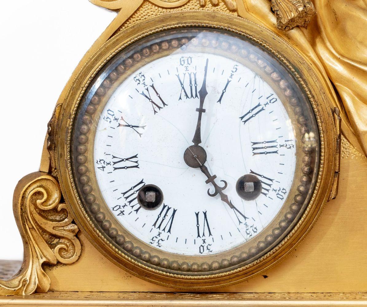 Restauration Gilt Metal Figural Mantel Clock, circa 1830, the circular enamel dial, with strike on bell movement, surmounted by a seated woman resting on a cushion, the rectangular base mounted with acanthus and foliate swags, on a scrolled base.