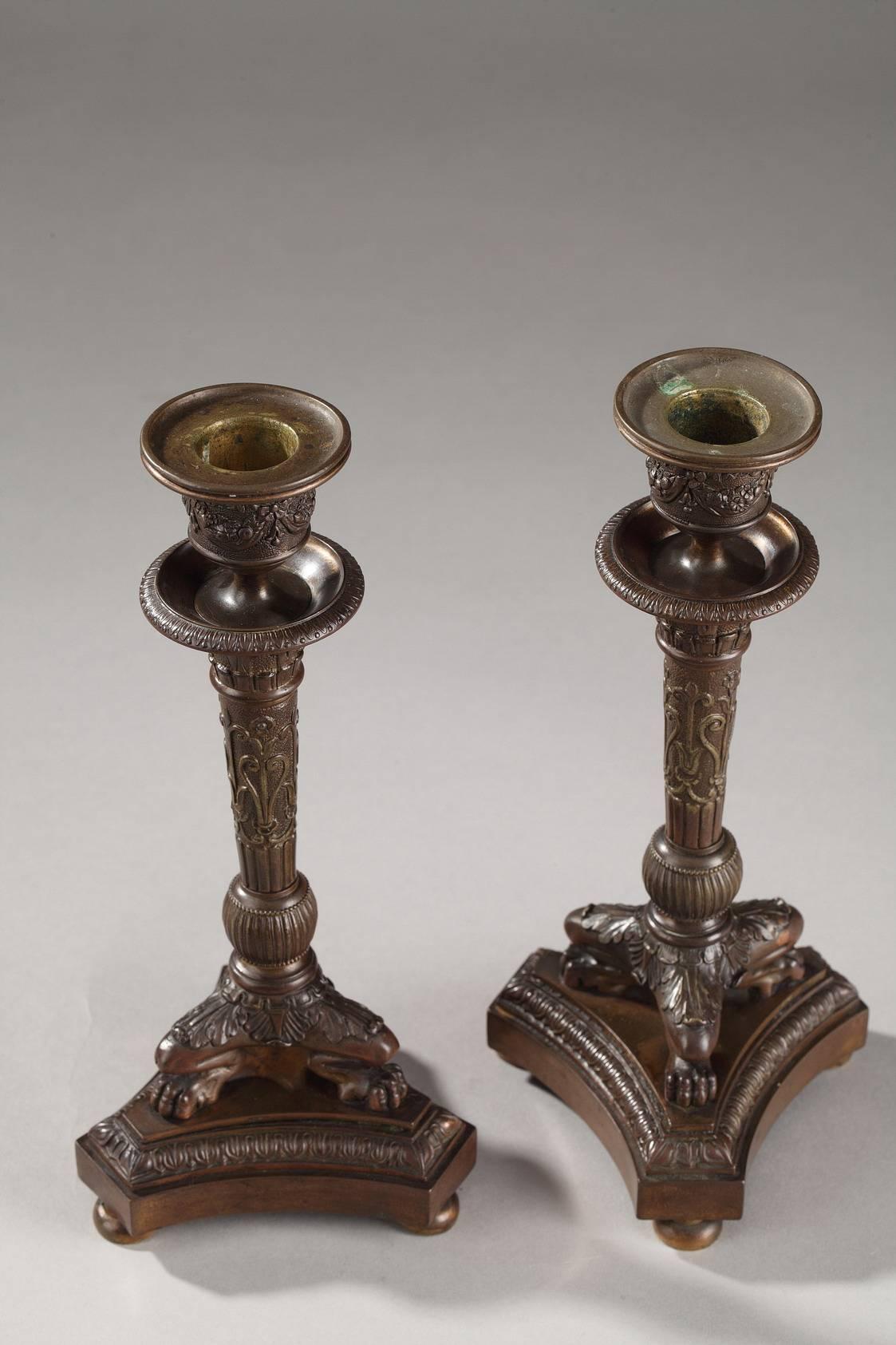 Coppia di piccoli candelieri in bronzo patinato che poggiano su tre zampe di leone impreziosite da palmette e foglie d'acanto. La base del treppiede è ornata da un motivo raie-de-coeur. Il fusto affusolato è scolpito con rami, scanalature, palmette