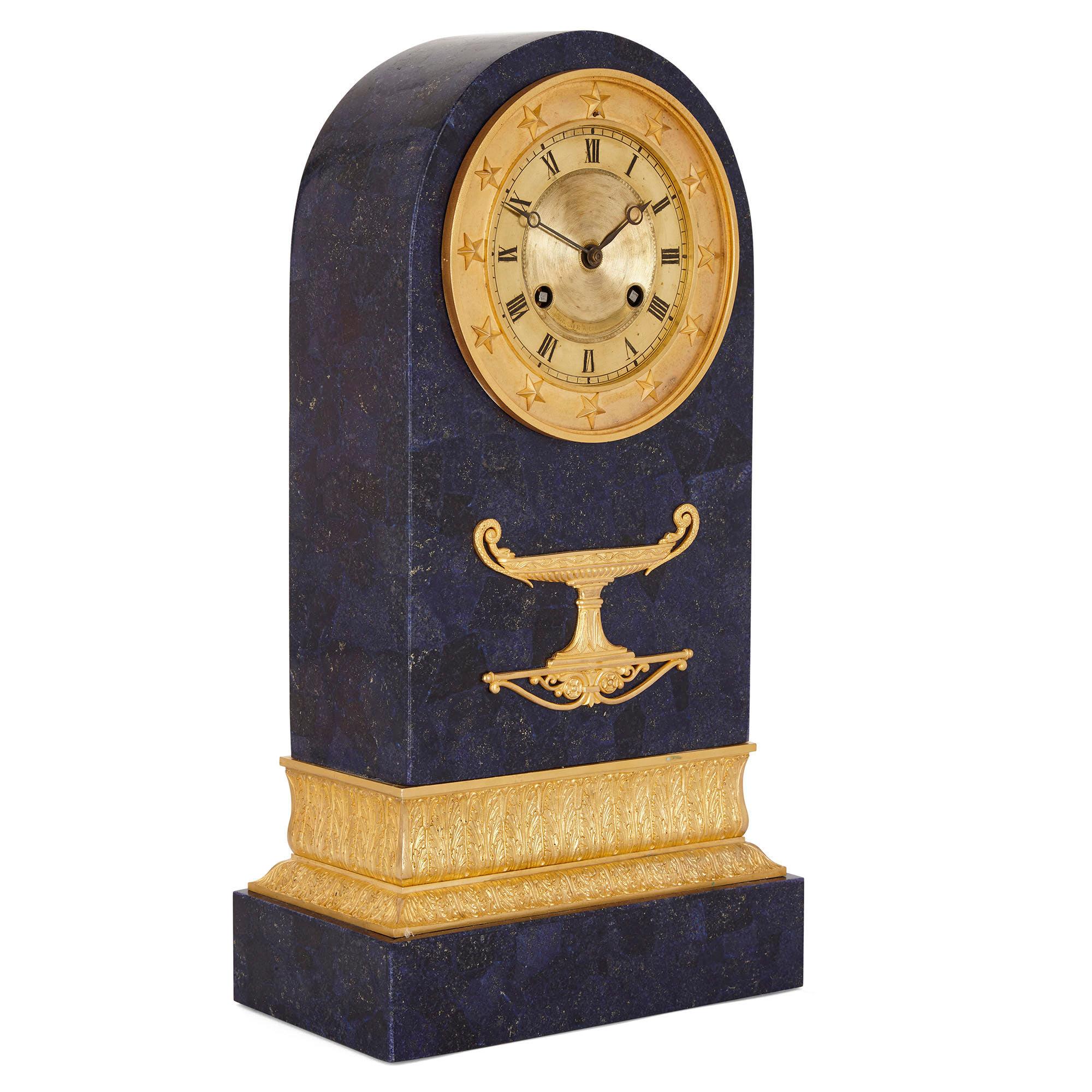 This mantel clock dates from the period of Charles X and is crafted from gilt bronze with a later lapis lazuli veneer. The clock body is columnar in form, having a rectangular profile with a semi-circular top. The body is supported by a gilt bronze