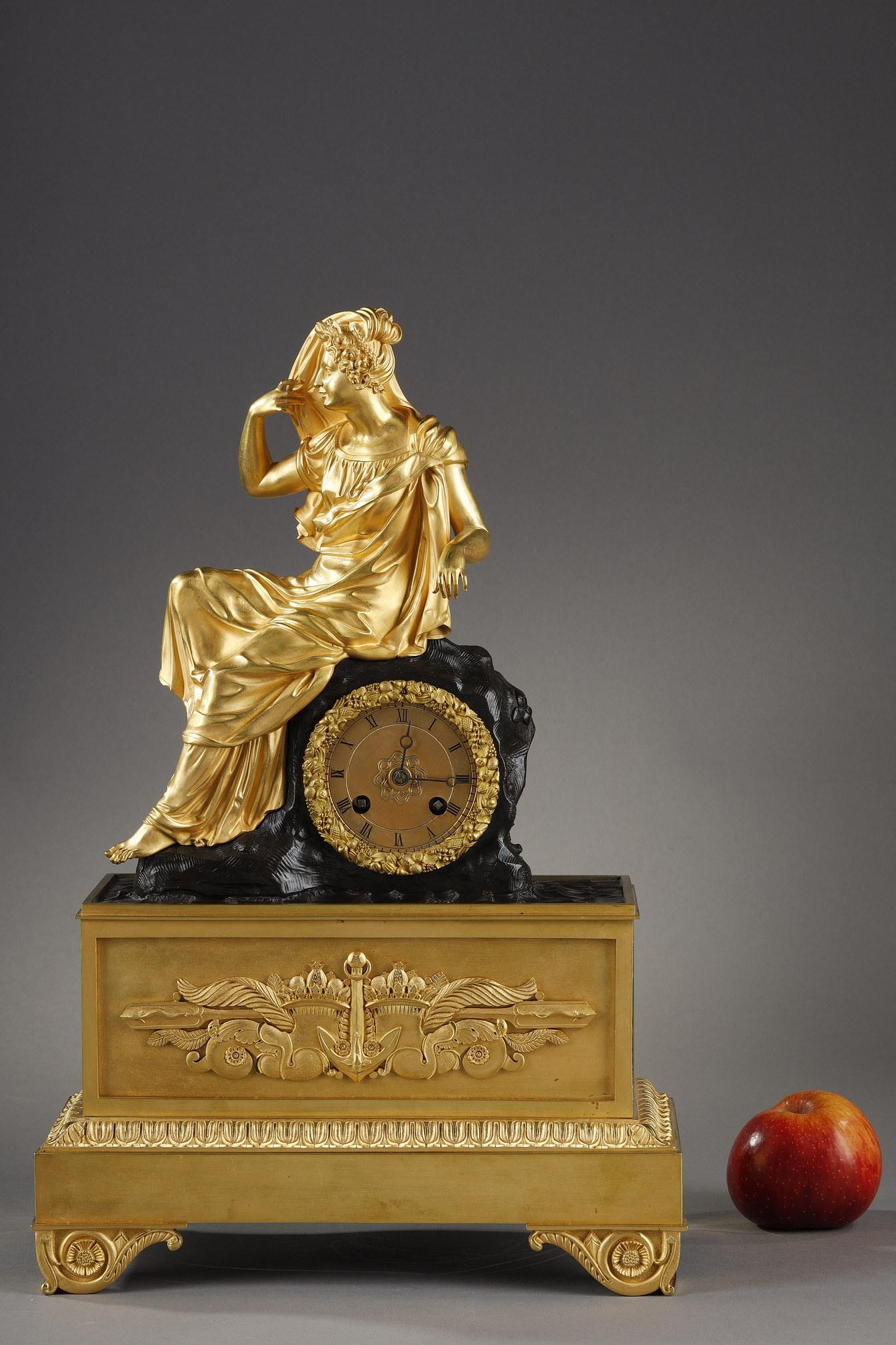 Clock in gilded and chiseled bronze representing a young woman sitting sideways dressed in the antique style, wearing a laurel crown and flowers, holding a veil above her head. She is seated on the dial set in a patinated bronze structure imitating