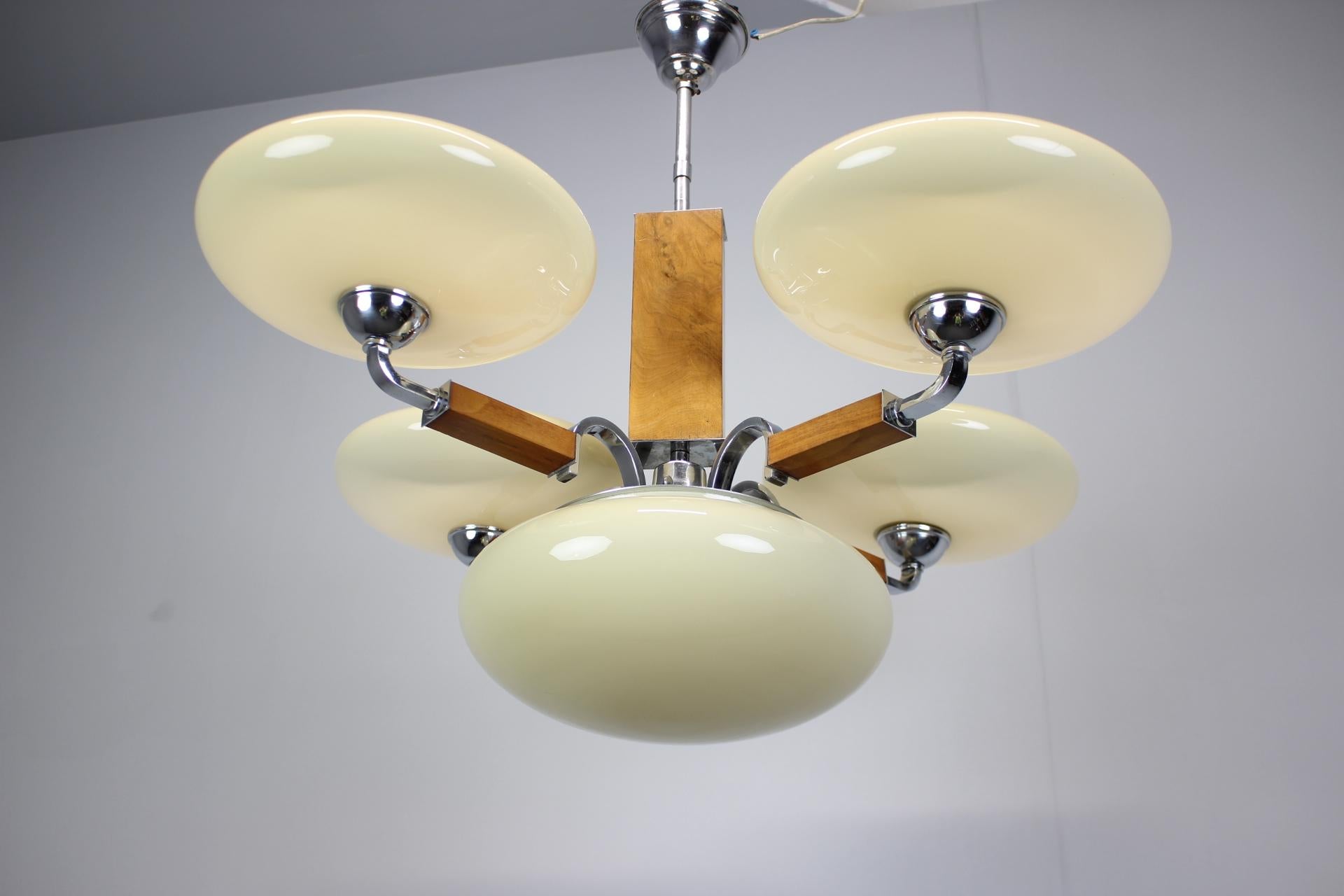 Esta elegante lámpara Art Decó se fabricó en la antigua Checoslovaquia en la década de 1930 y muestra una refinada combinación de cromo, madera de tonos cálidos y pantallas de cristal crema suavemente curvadas. La estructura de cinco luces encarna