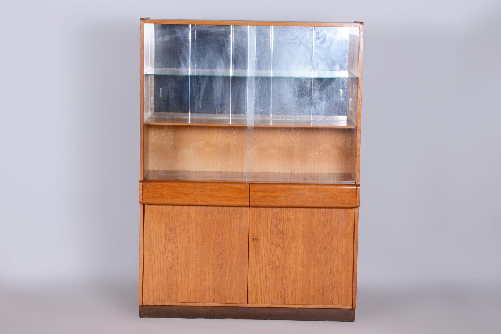 Esta vitrina de los años 40 muestra una elegante mezcla de estilos Art Decó y de mediados de siglo, característicos de la Checoslovaquia de posguerra. Producido por el renombrado fabricante Jitona Soběslav, está elaborado con madera de roble de alta