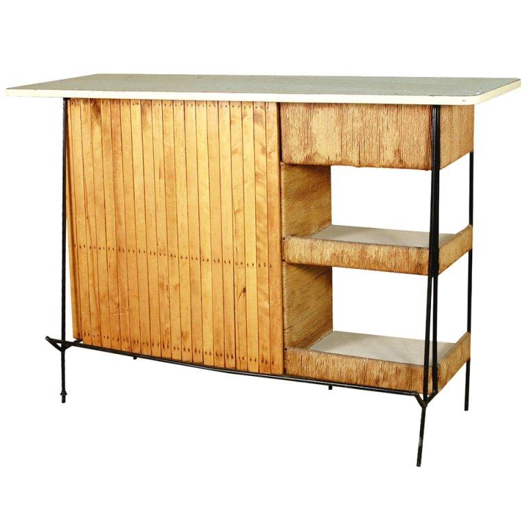 This Arthur Umanoff Mid-Century Modern cocktail bar features cut-out side windows for storage and keeping barware and spirits on display. Made of oak slats with paper cording and woven wicker accents on a wrought iron base; Formica top and shelf