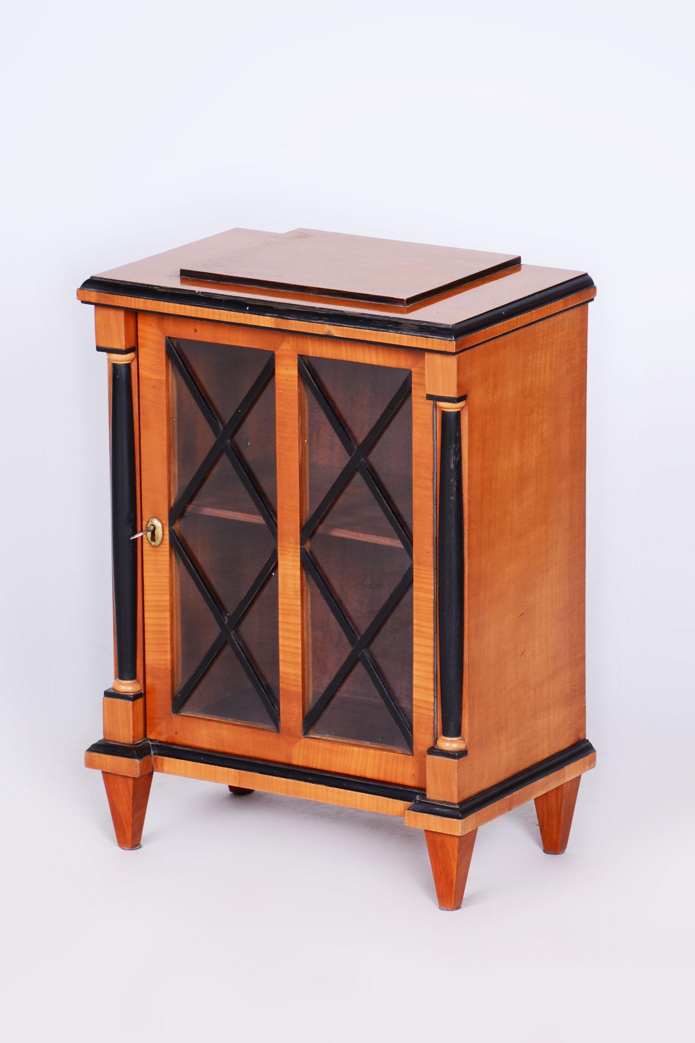 This elegant small Biedermeier display cabinet comes from Czechoslovakia from the mid-19th century. It is made of solid wood with cherry veneer and features ebonized columns and decorative moldings. The cabinet has undergone careful restoration and