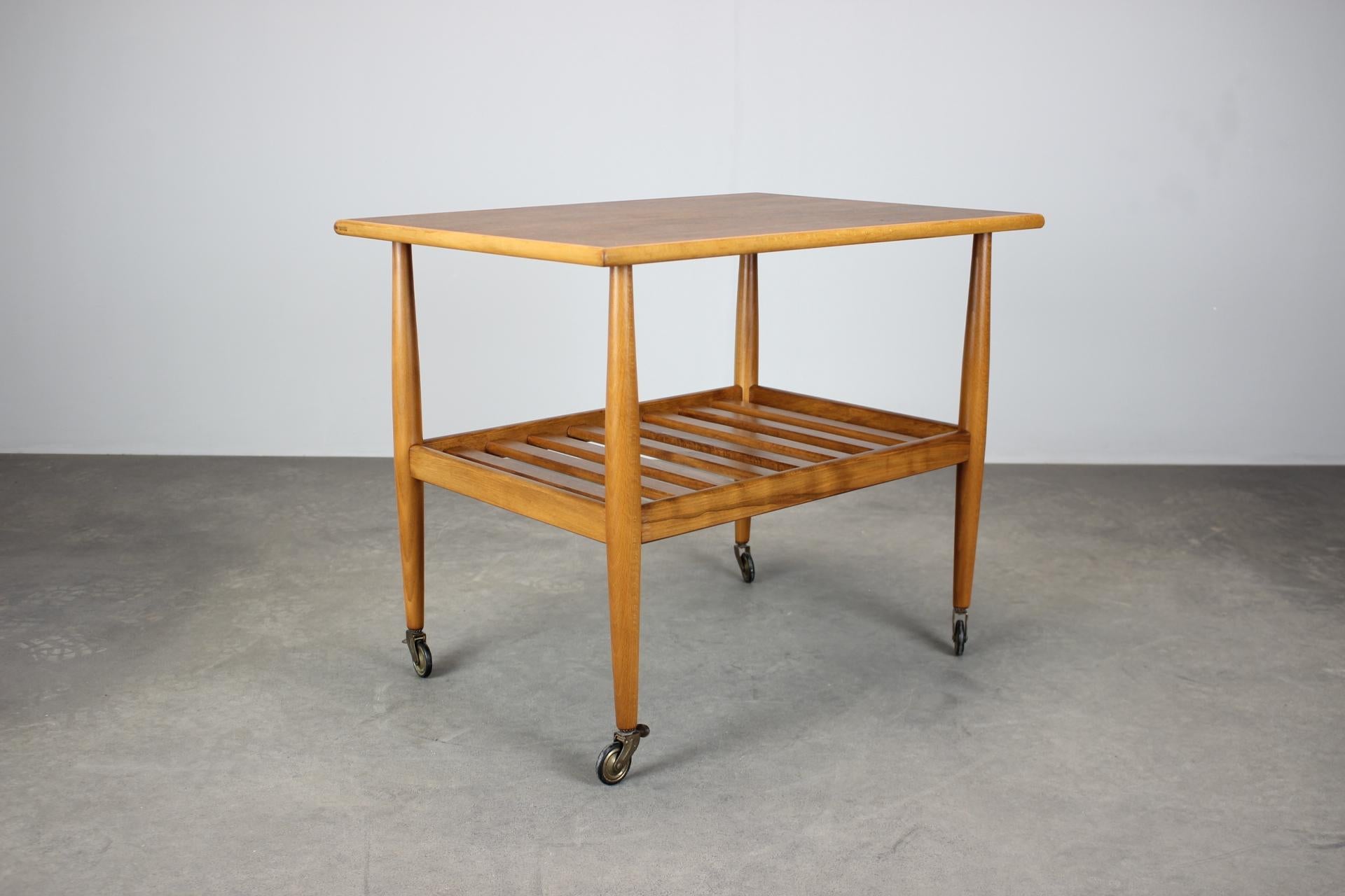 Esta mesa rodante danesa de mediados de siglo, de la década de 1960, tiene tablero chapado en teca, estante inferior de tablillas y ruedas originales, una de ellas con freno. Un diseño versátil y compacto adecuado como mesa de servicio, mesa