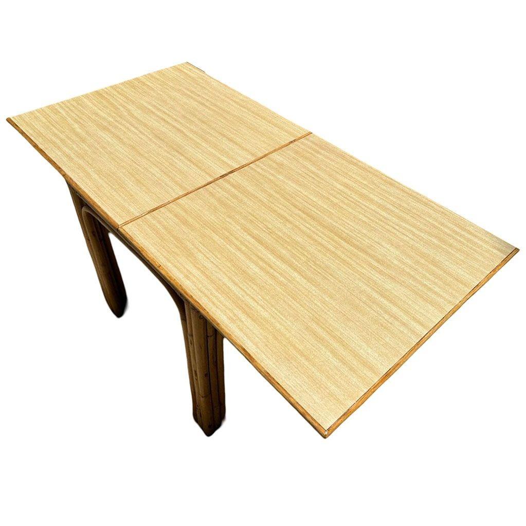 Esta mesa de comedor de ratán y fórmica veteada de madera de mediados de siglo, bellamente restaurada e inspirada en Paul Frankl, tiene un tablero de 2 hojas articuladas expansibles de color rubio, que permite ampliarla fácilmente para acomodar a