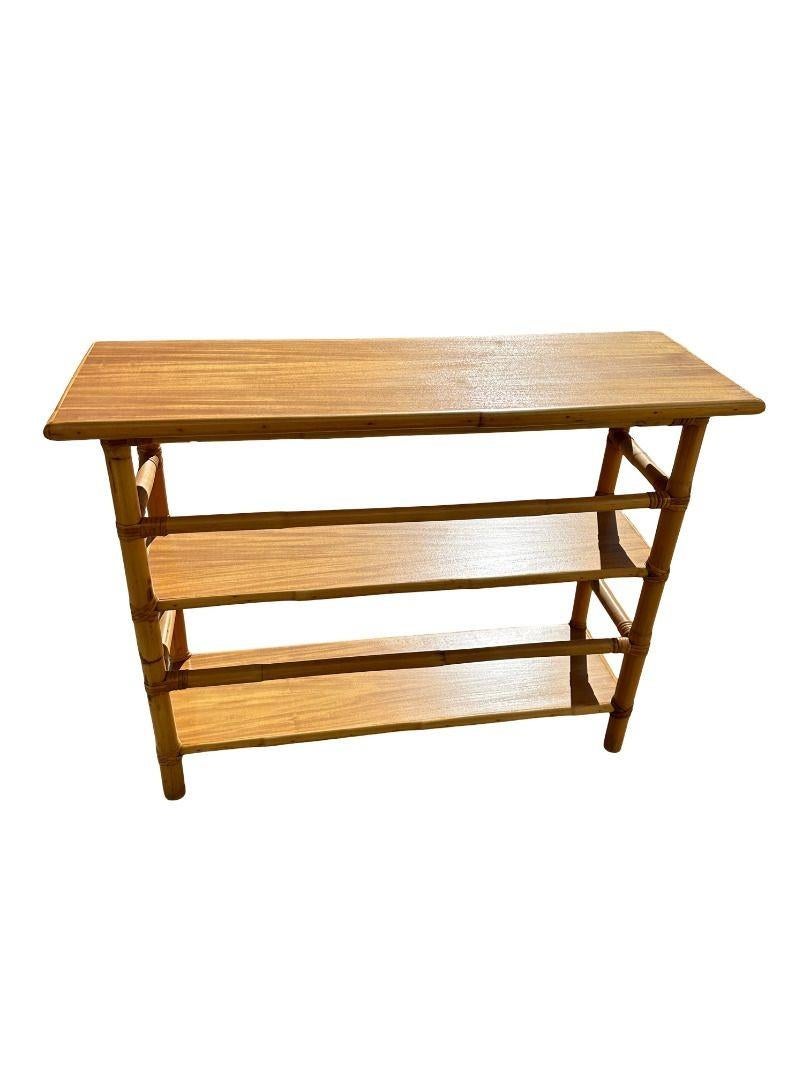 Postwar three-tier console table Bookshelf with mahogany tops and a wrapped stick rattan frame. The table has mahogany tops along each shelf which are decorated with rattan trim and each shelf has rattan pole rails for holding books in place.

This