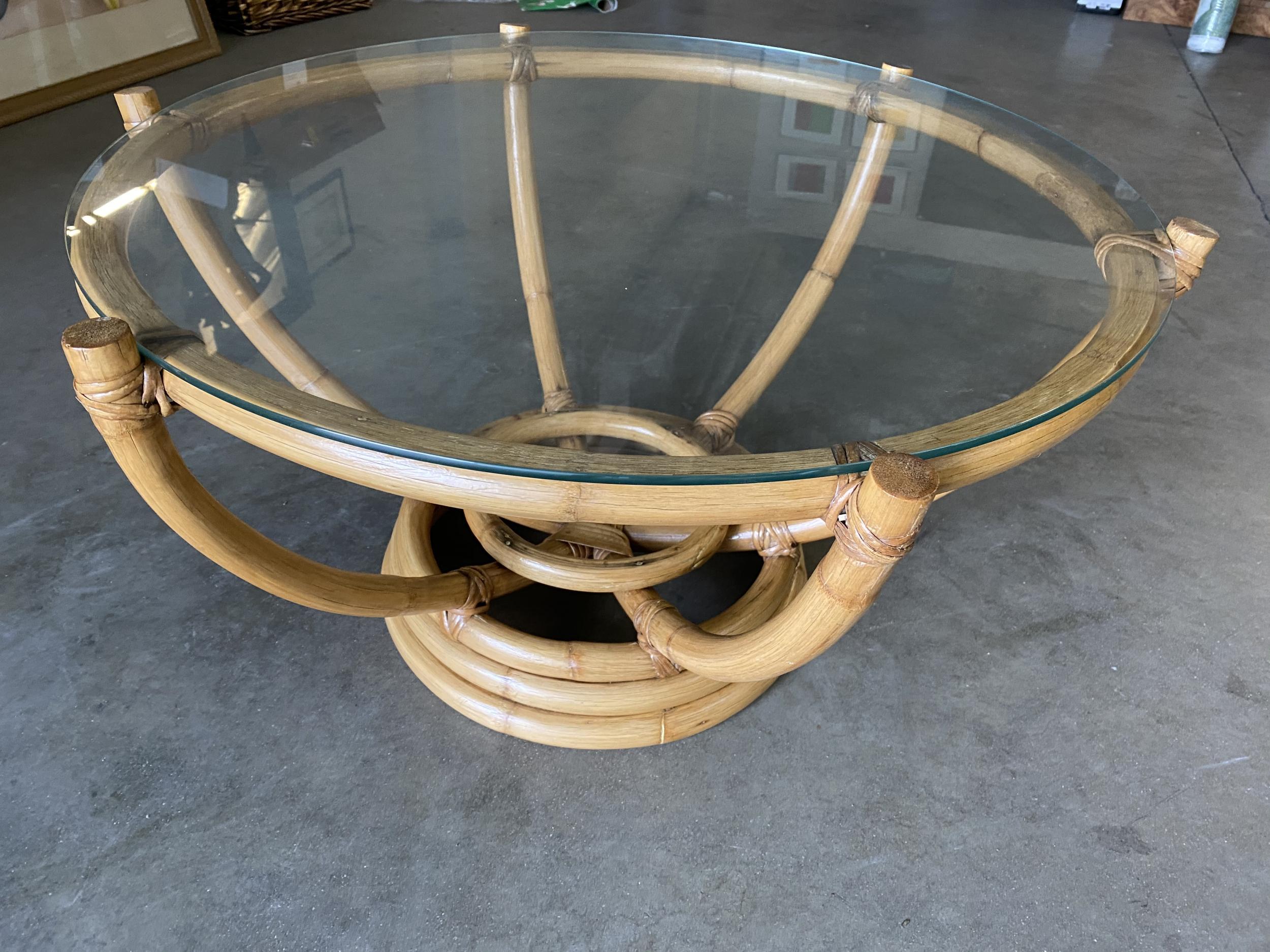 Made circa 1940, this six-pole rattan coffee table has an unusual stacked rattan round base. The six rattan poles organically branch outward to a circular top made of a single piece of bent rattan. The glass top rests atop the poles, exuding an