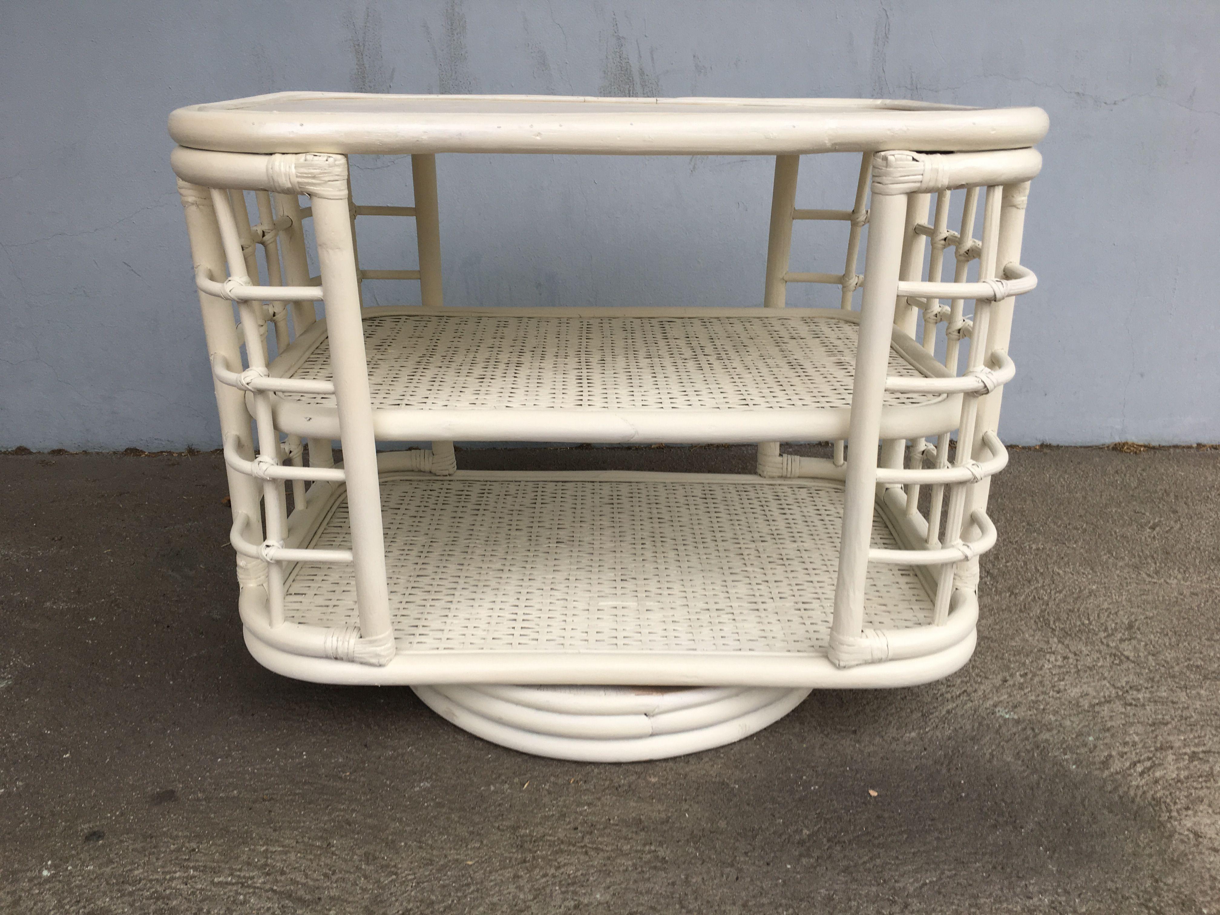 White painted midcentury era rattan side table bookshelf featuring two shelves with skeleton side walls and a swivel base for easy book location. Restored to new for you. All rattan, bamboo and wicker furniture has been painstakingly refurbished to