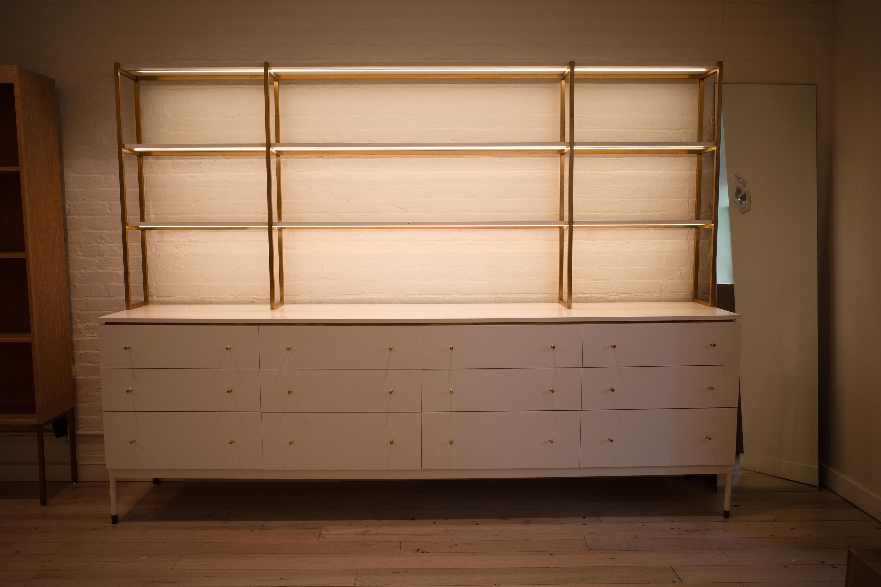 This custom-built hutch provides both storage and display space. It features a steel frame with wood in a white lacquer finish and is accented with brass hardware and detailing. The unit is equipped with LED track lighting to highlight items on its