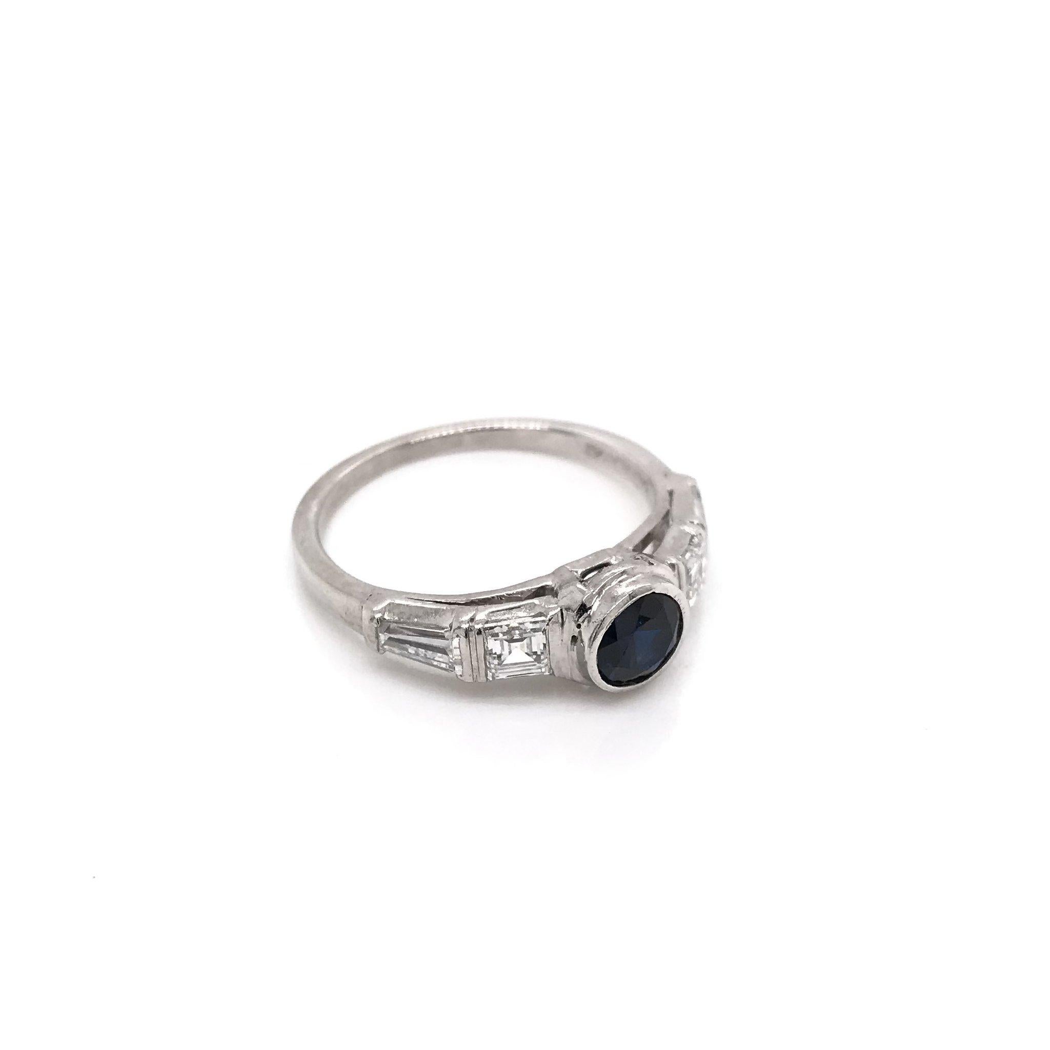 This charming Mid Century piece was crafted sometime during the Retro design period ( 1940-1960 ). The center stone is a darkly hued blue sapphire measuring approximately 0.57 carats. The blue sapphire is bezel set and flanked by two Asscher cut